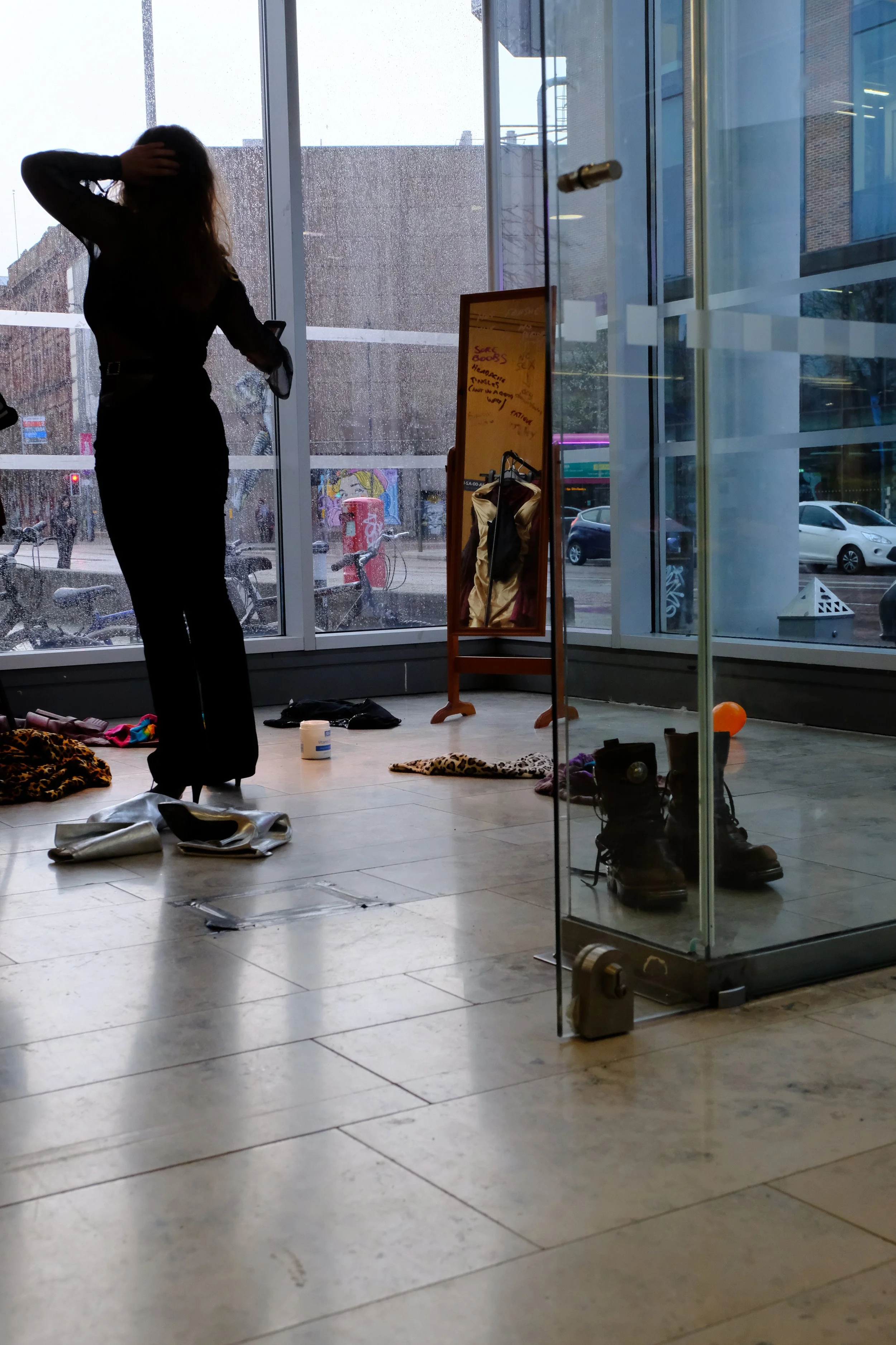 A woman with long hair wearing black clothing is standing inside a store near large glass windows with rain outside. There's a mirror, shoes, apparel, and miscellaneous items on the floor, and a reflection of the street is visible through the window.