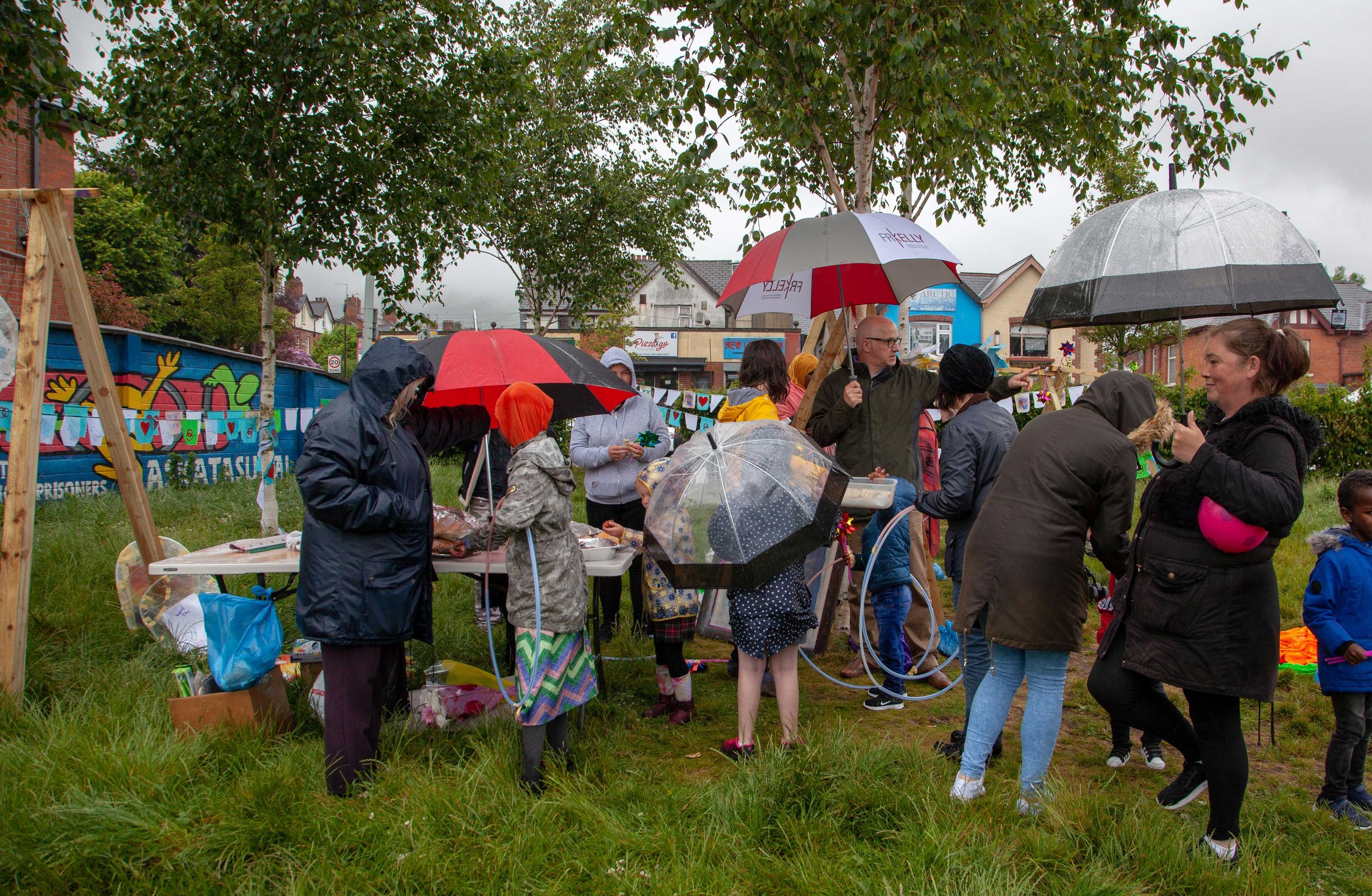 People attending an outdoor community event on a rainy day, holding umbrellas, standing around tables, with decorations and a colorful mural on a fence in the background.