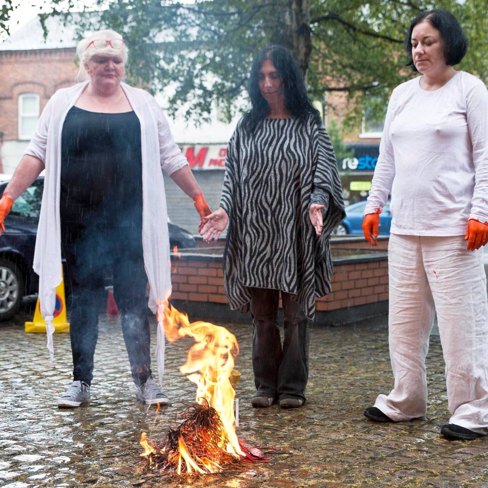 Three women stand outdoors around a small fire on a cobblestone ground, holding hands with head bowed and eyes closed, with trees and buildings in the background. The woman on the left has white hair, wears glasses on her head, a white cardigan, black shirt, and blue jeans. The woman in the center has long black hair, is wearing a zebra-patterned dress, and appears to be praying. The woman on the right has short black hair, is dressed in a white outfit, and wears orange gloves.