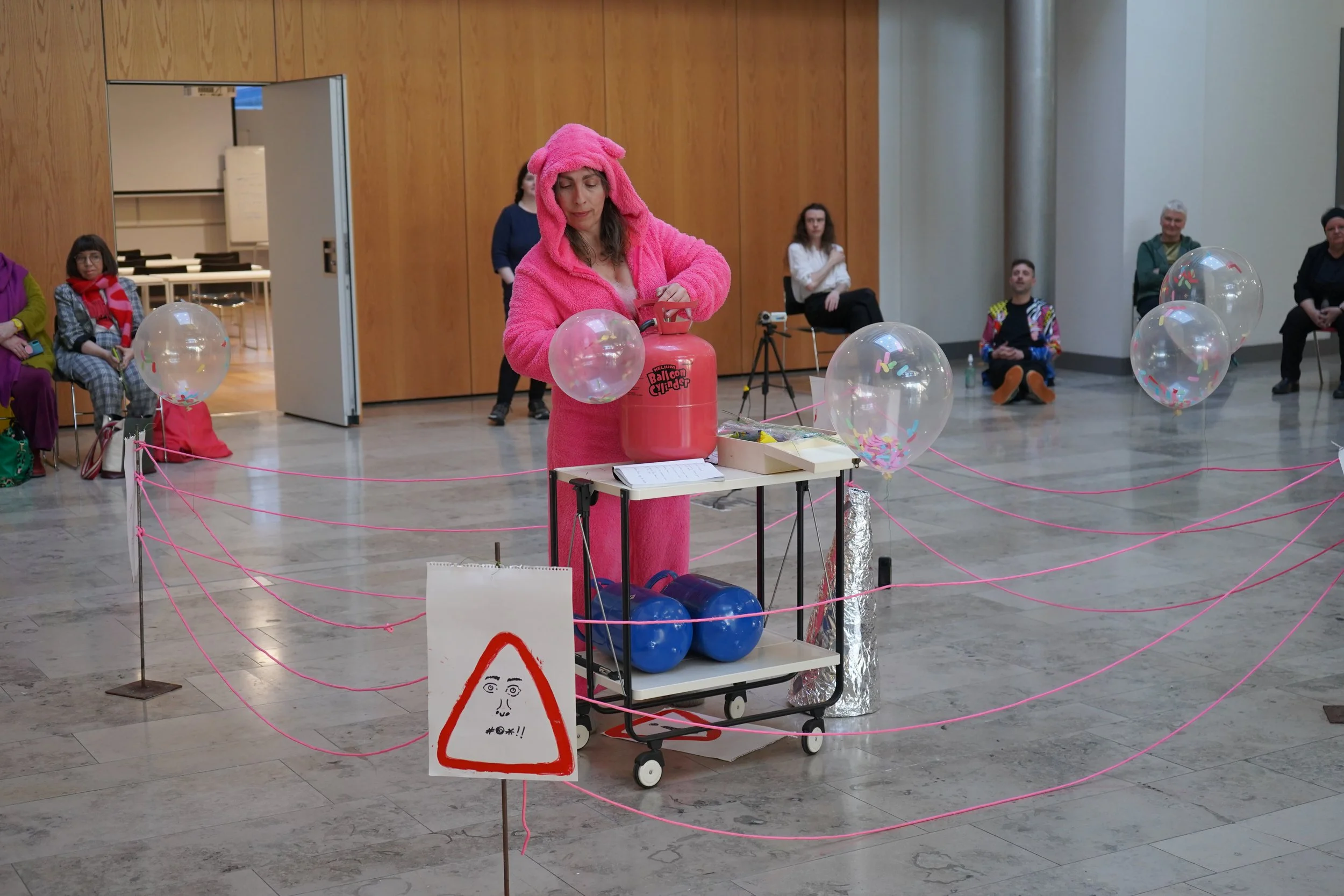 Woman in a pink costume with a hood that resembles a cartoon animal, operating a helium balloon cylinder at an indoor event. The scene is enclosed with pink strings and balloons filled with confetti, with seated and standing people watching in the ba