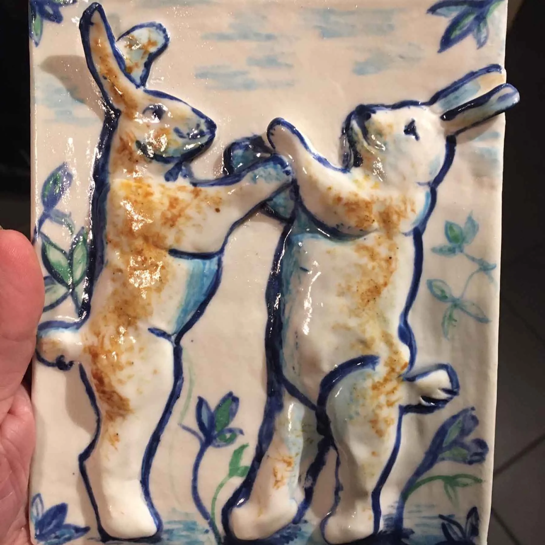 Ceramic tile featuring painted line drawings of two rabbits, outlined in blue with orange accents, surrounded by green leaves and blue decorative elements.