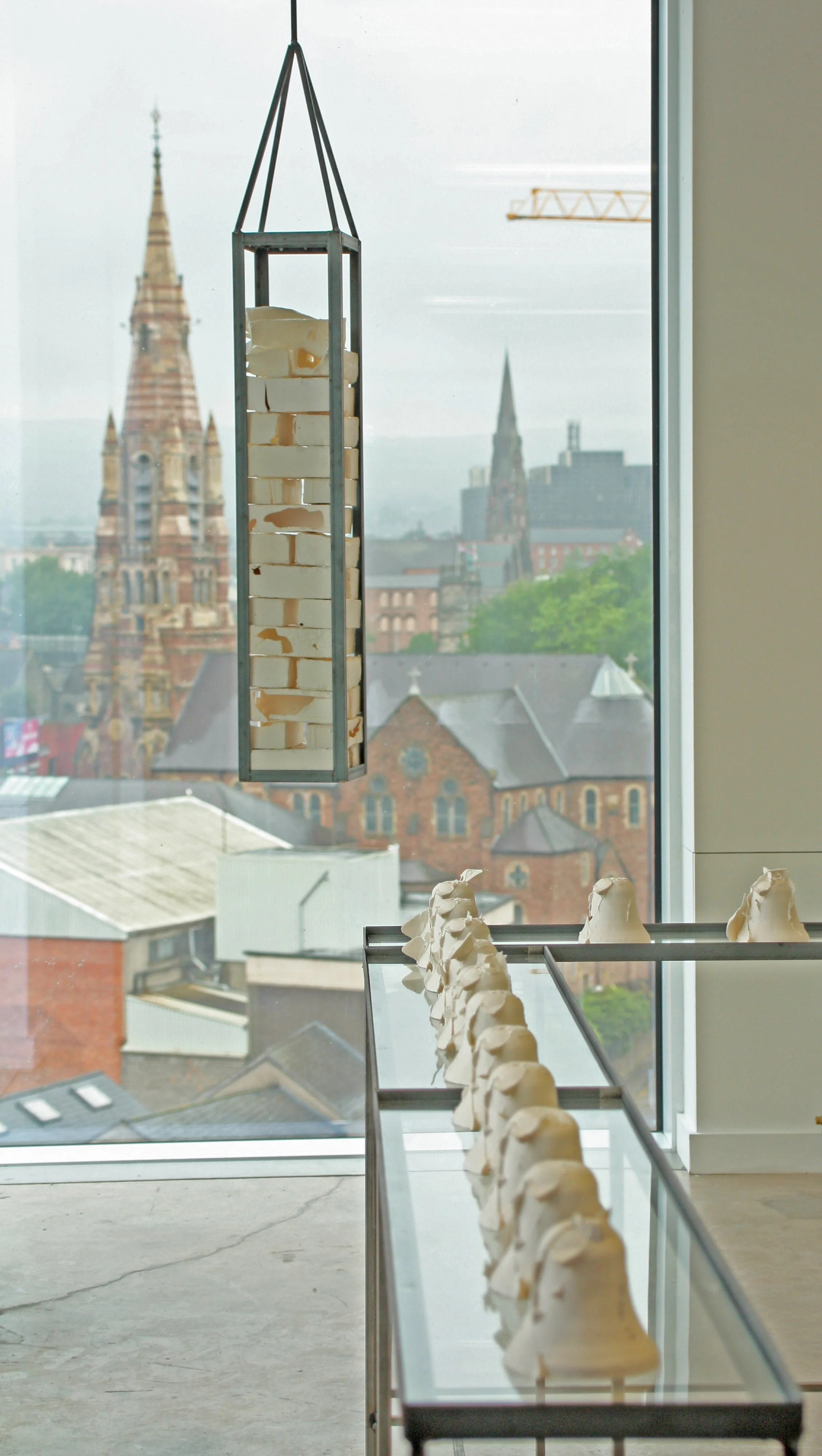 Sculpture of a stack of white porcelain bricks inside a metal frame hanging in front of a large window with cityscape view.