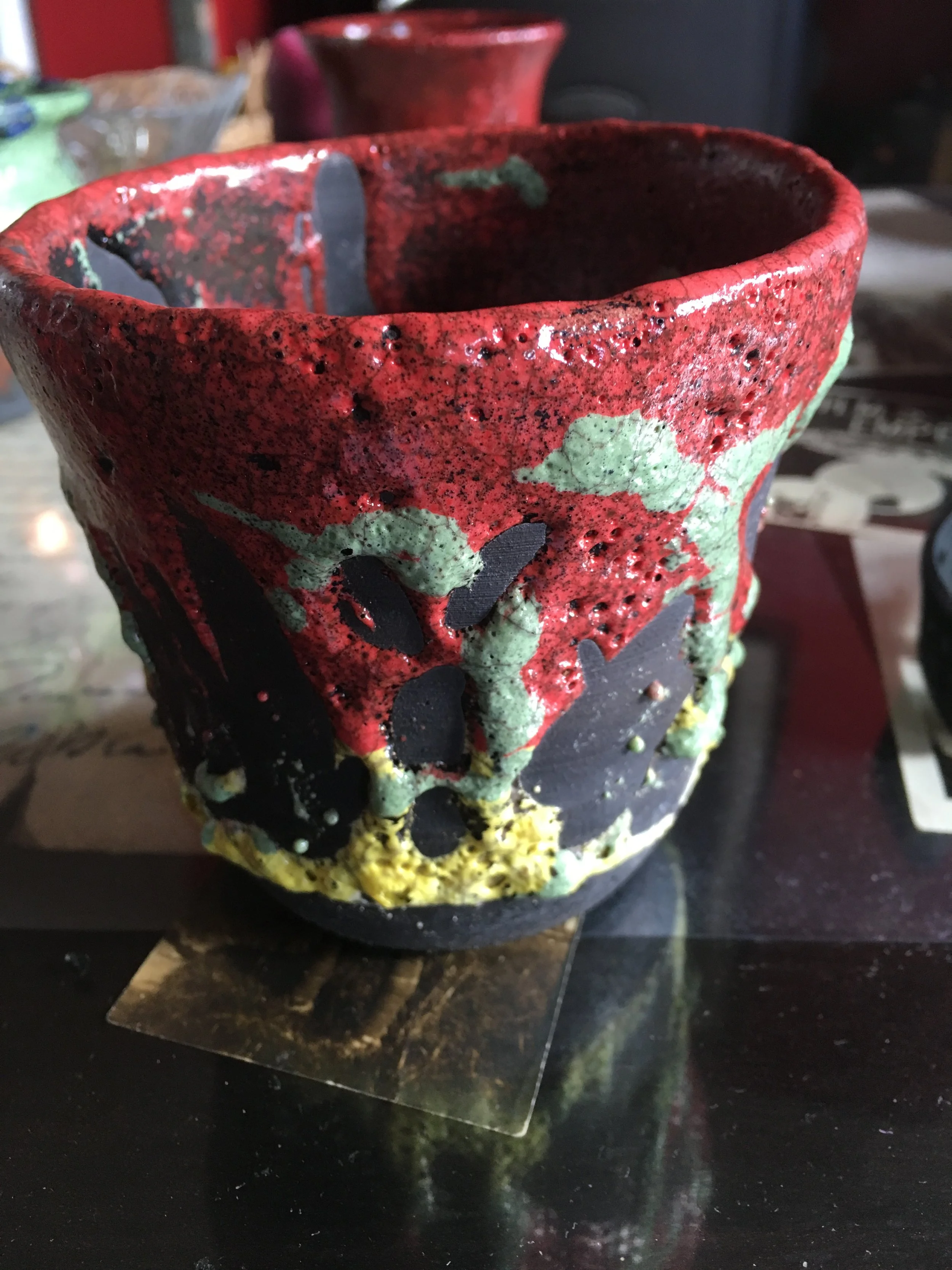 Close-up of a colorful ceramic vase with a textured surface, decorated with red, green, black, and yellow glaze, and featuring cut-out patterns. The vase is placed on a dark surface with other decorative items blurred in the background.