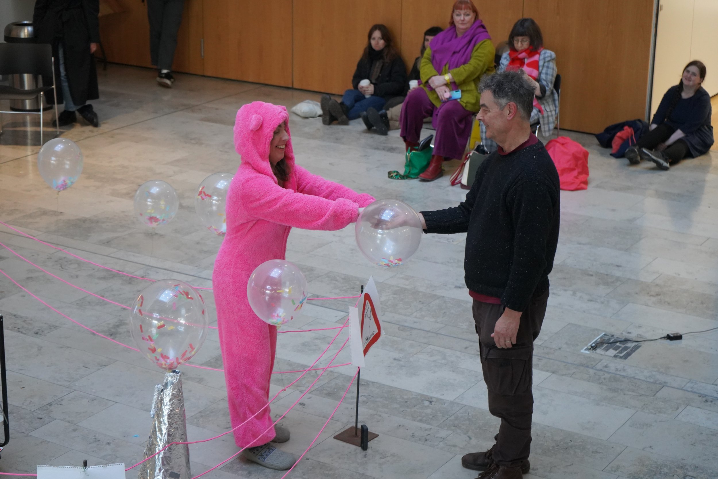 A woman dressed in a pink fluffy animal costume, possibly resembling a bear, is holding hands with a man in a black sweater as they stand inside a room decorated with pink ribbons, balloons filled with confetti, and a small sign on a table. Several people are sitting on the floor and chairs in the background, watching and smiling.