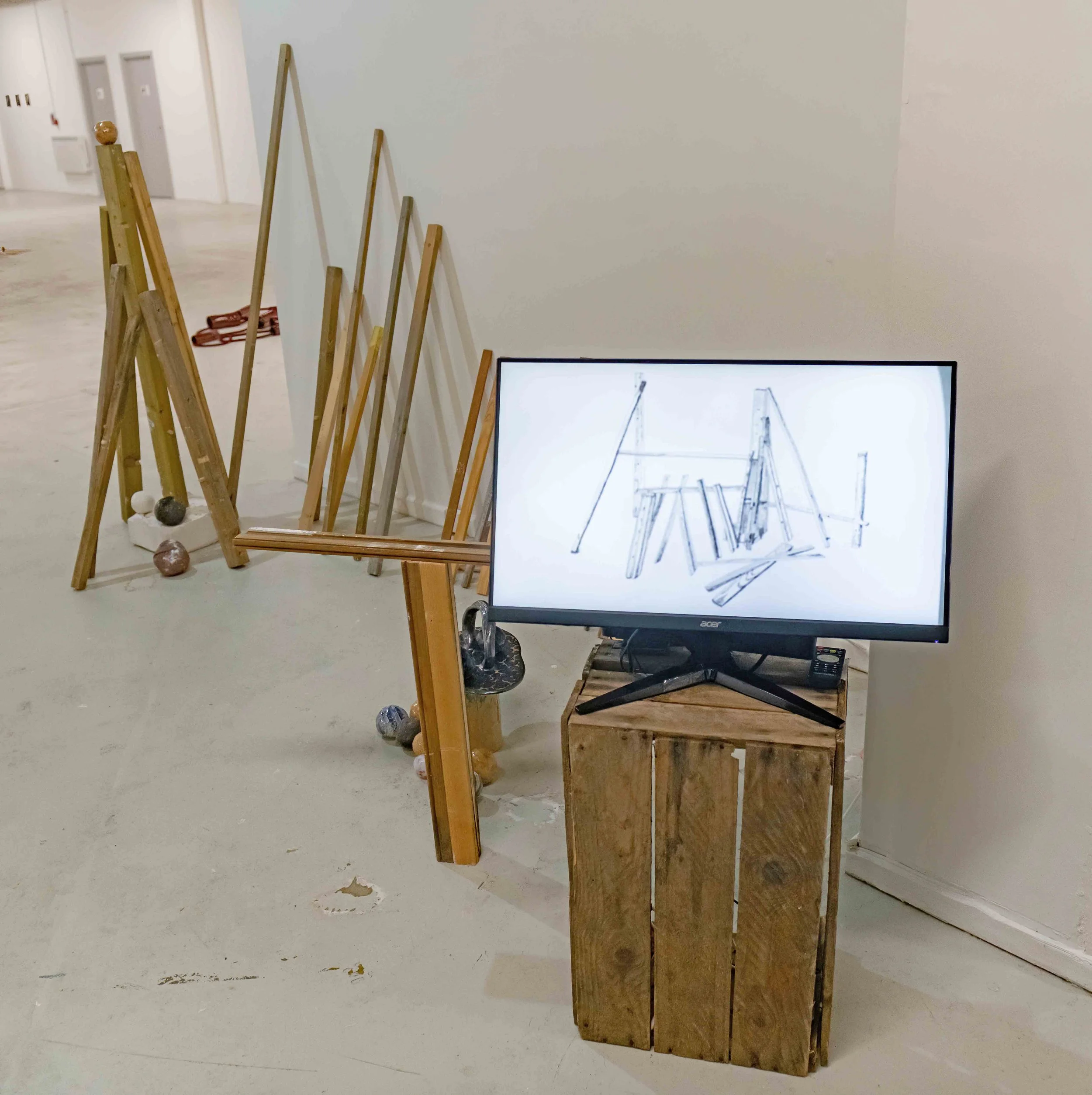 Wooden sticks arranged in a diagonal line in front of a large white wall, with a computer monitor displaying a line drawing of chairs and a staircase. The room has a light-colored floor and minimalist decor.