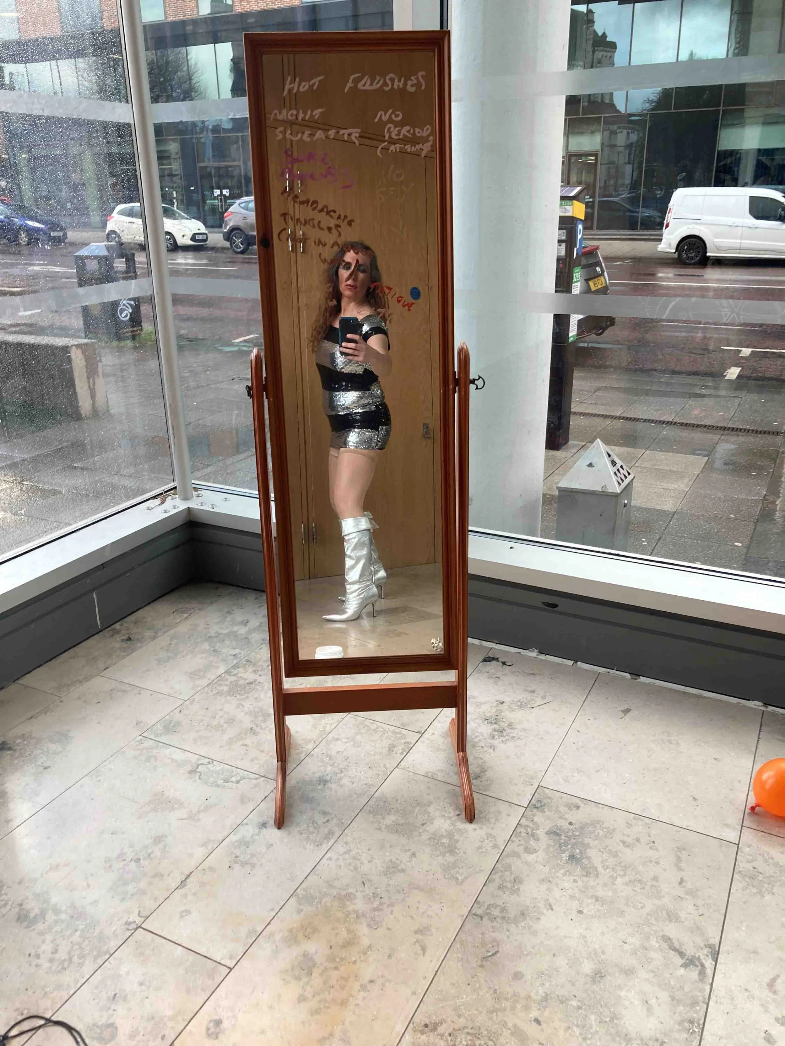A woman taking a selfie in a full-length mirror in a modern indoor space with large windows showing a rainy street outside. She has long brown hair, is wearing a shiny silver and black dress, and knee-high silver boots.
