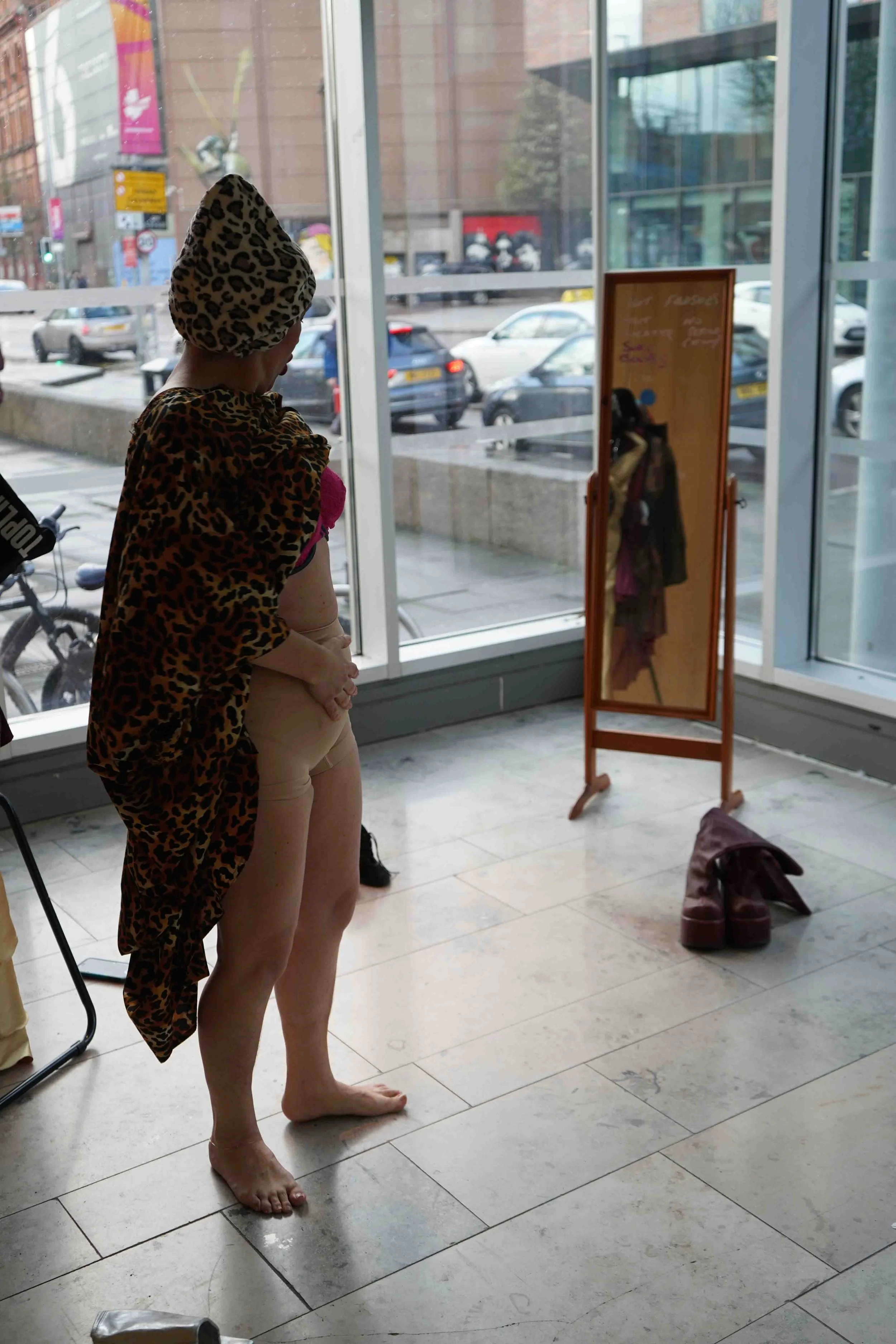 A woman stands barefoot indoors, wearing a leopard print headscarf and matching shawl, and beige shorts, looking at a standing mirror.