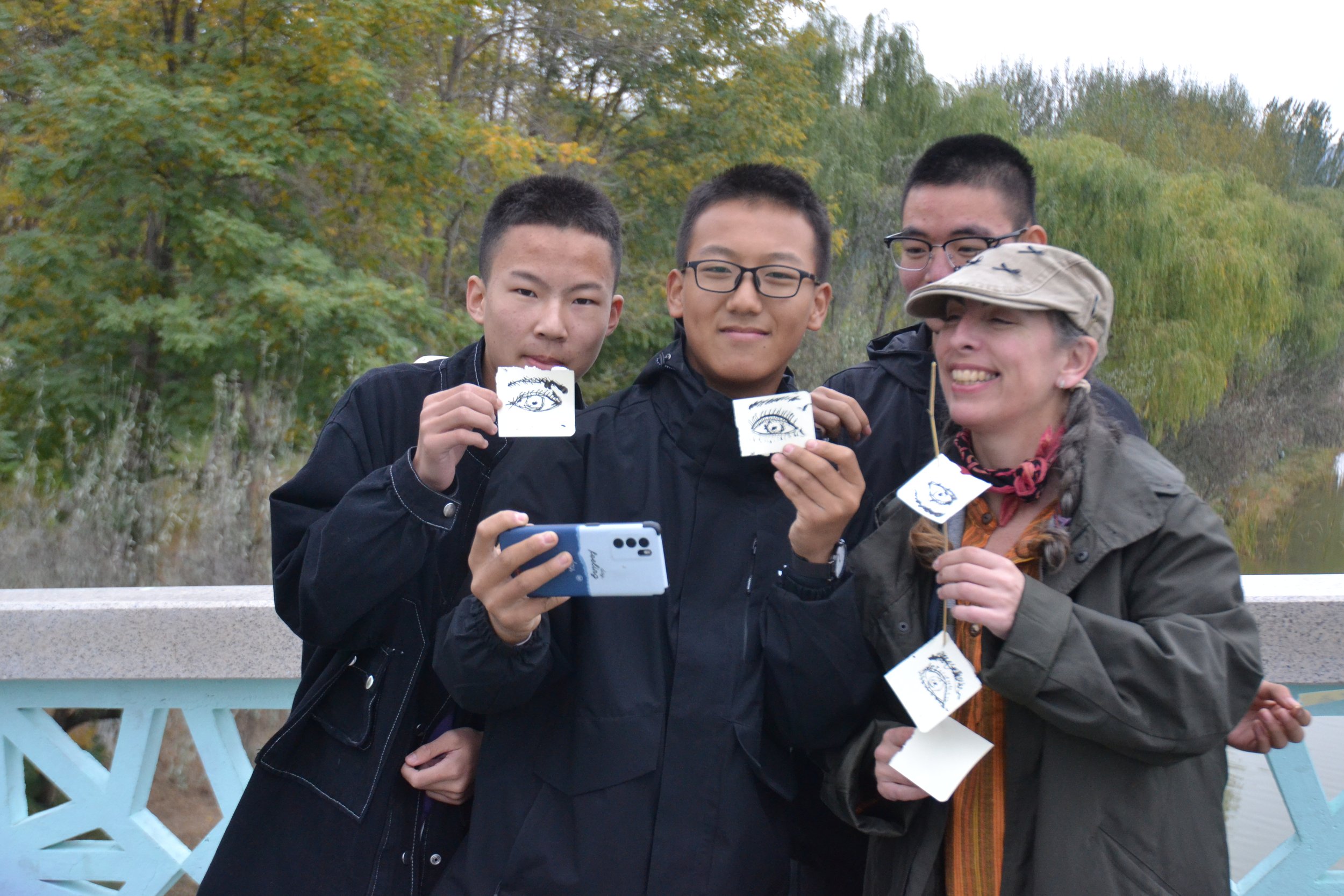 A group of four people outdoors holding small illustrated pieces of paper with eye drawings, smiling, with trees in the background.