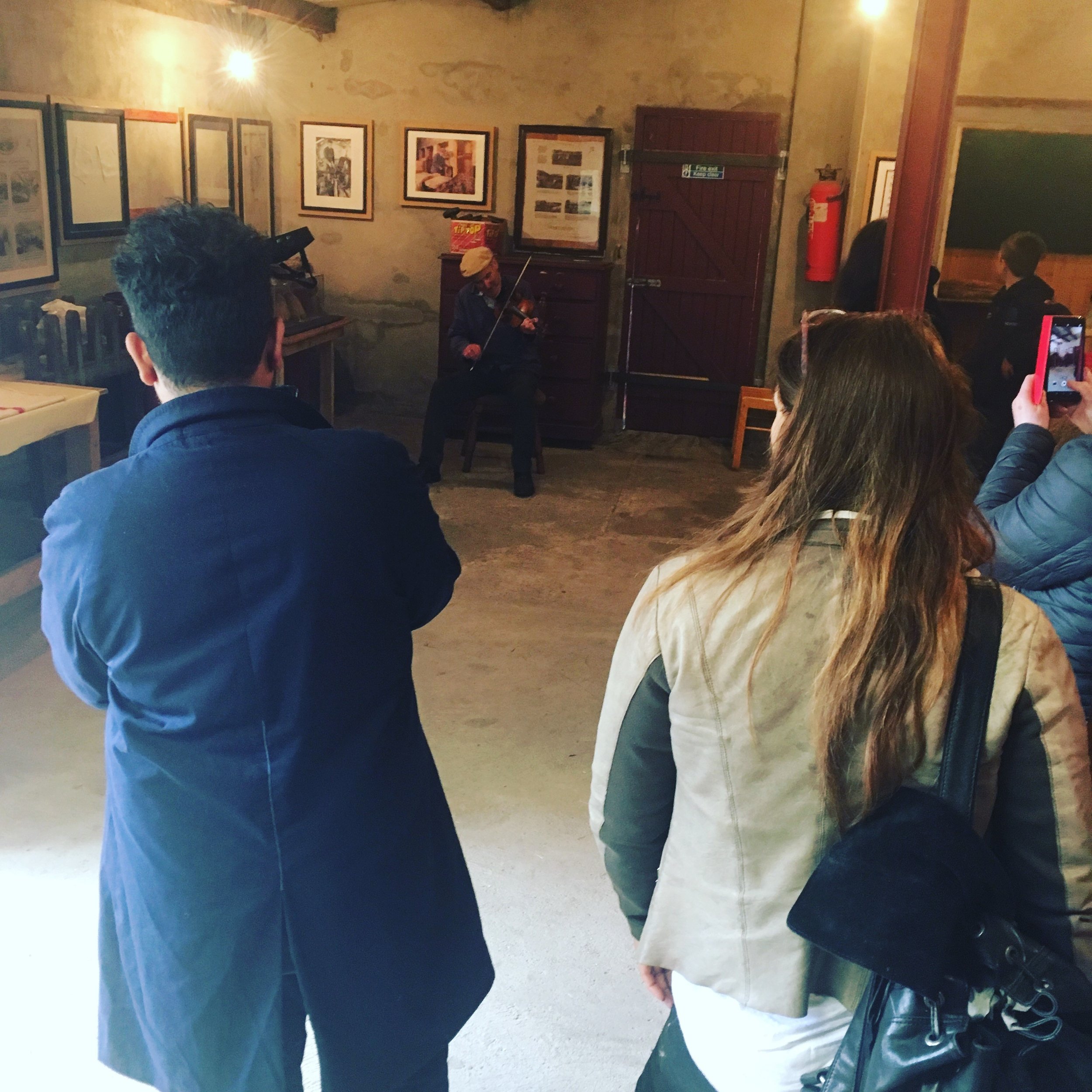 A group of people sitting indoors, facing a performer playing the violin. The audience members are diverse, with some holding phones. The room has framed pictures on the wall, a wooden door, and a fire extinguisher.