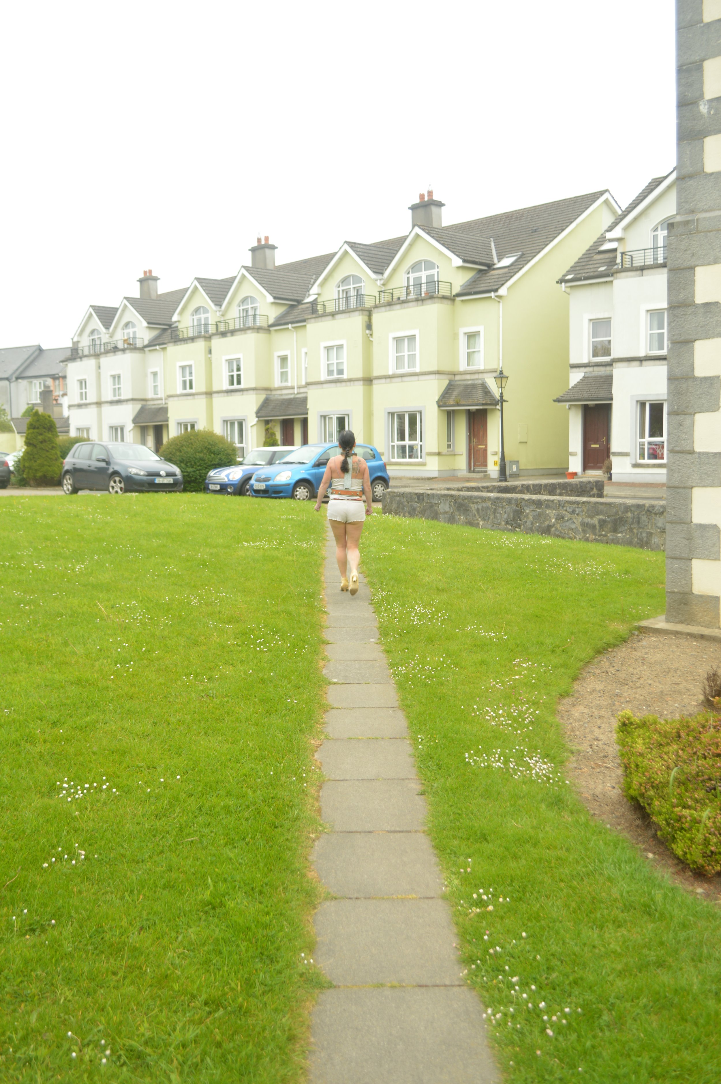 A woman walking on a sidewalk through a grassy area toward a row of light-colored suburban houses with cars parked in front.