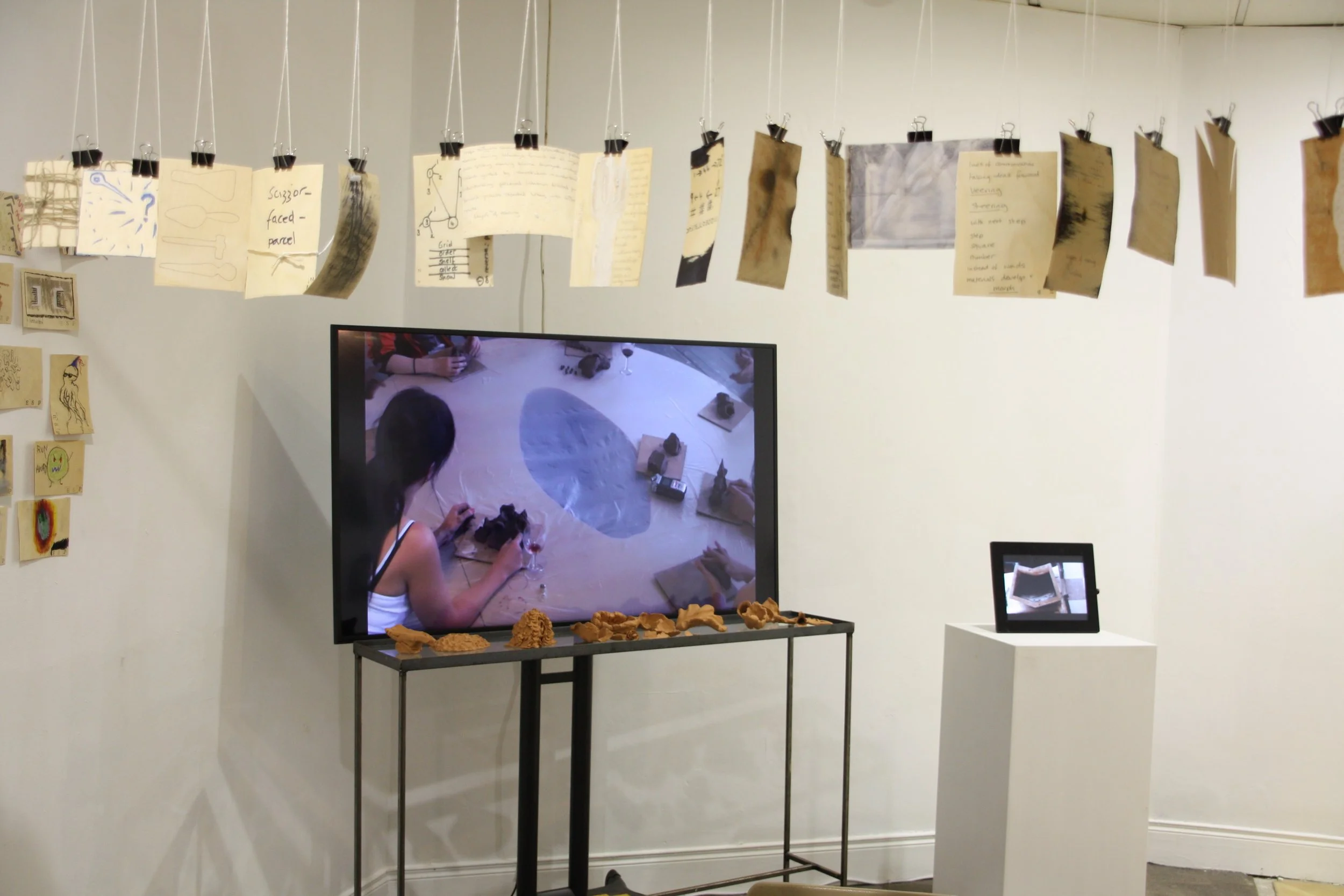 An indoor art exhibit features a string of pages or sketches hanging from the ceiling, with a TV and smaller display screen showing the same installation scene. Decorative objects are on a table in front of the TV.