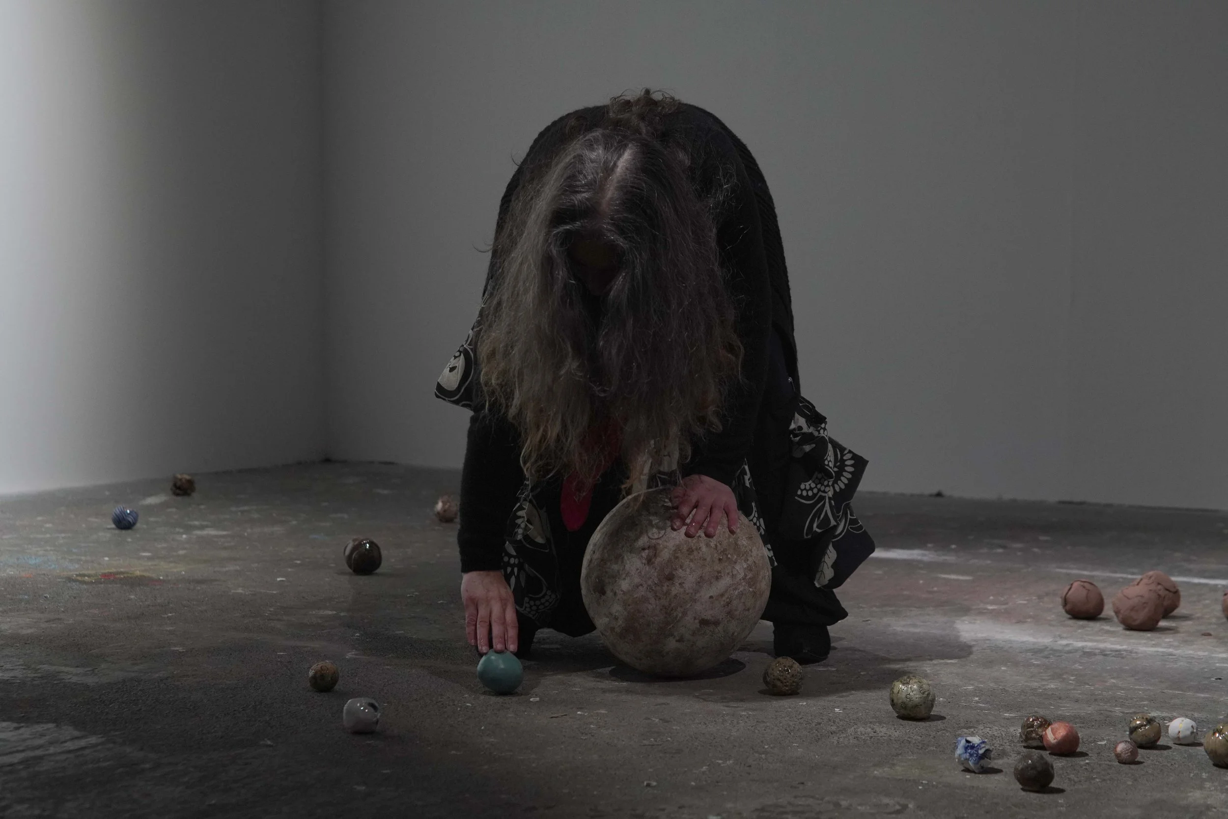 A person with long hair leaning over a large spherical object. They are surrounded by smaller spherical objects scattered on the floor in an empty room with gray walls.