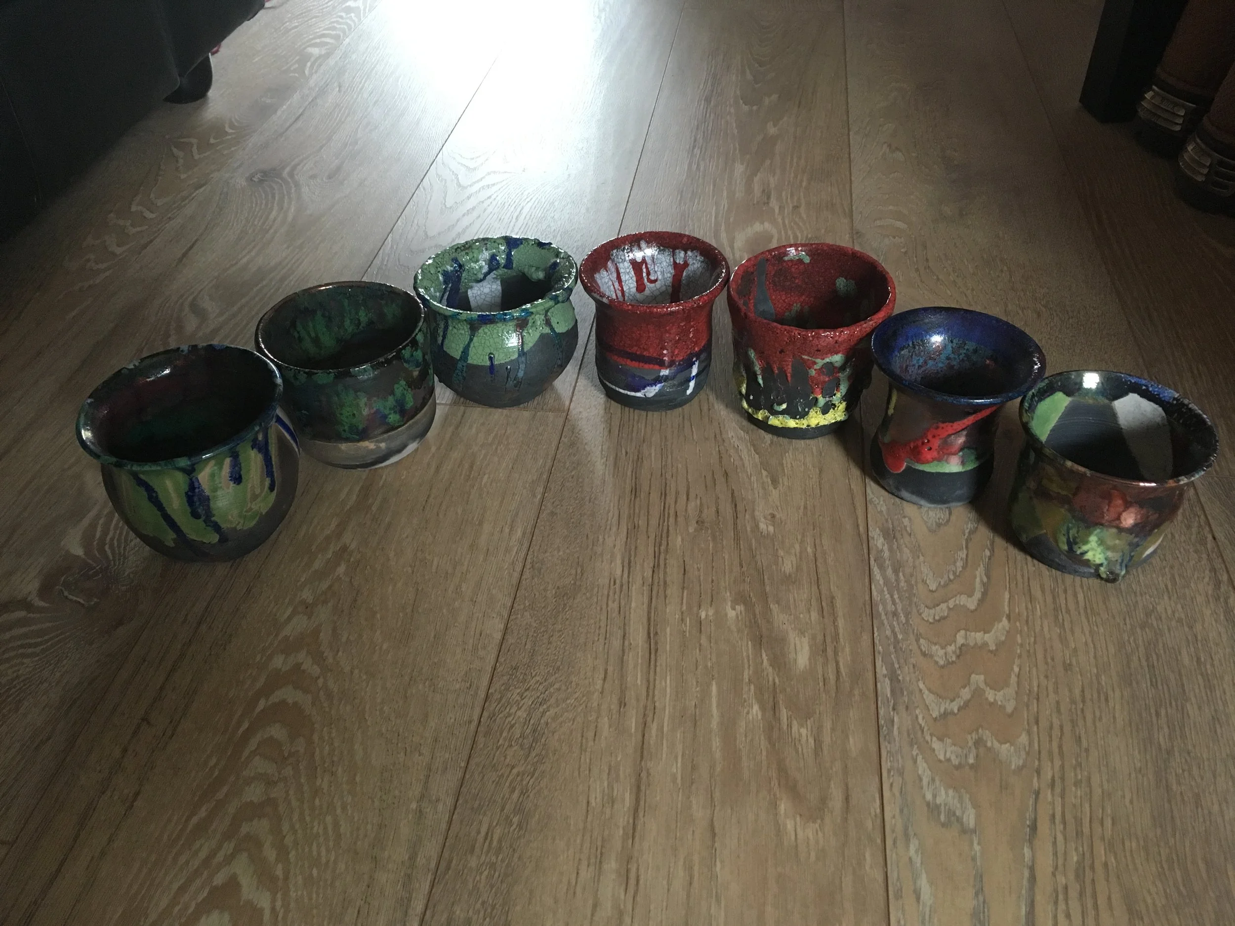 Six colorful ceramic cups arranged in a line on a wooden floor with sunlight illuminating the top part of the image.