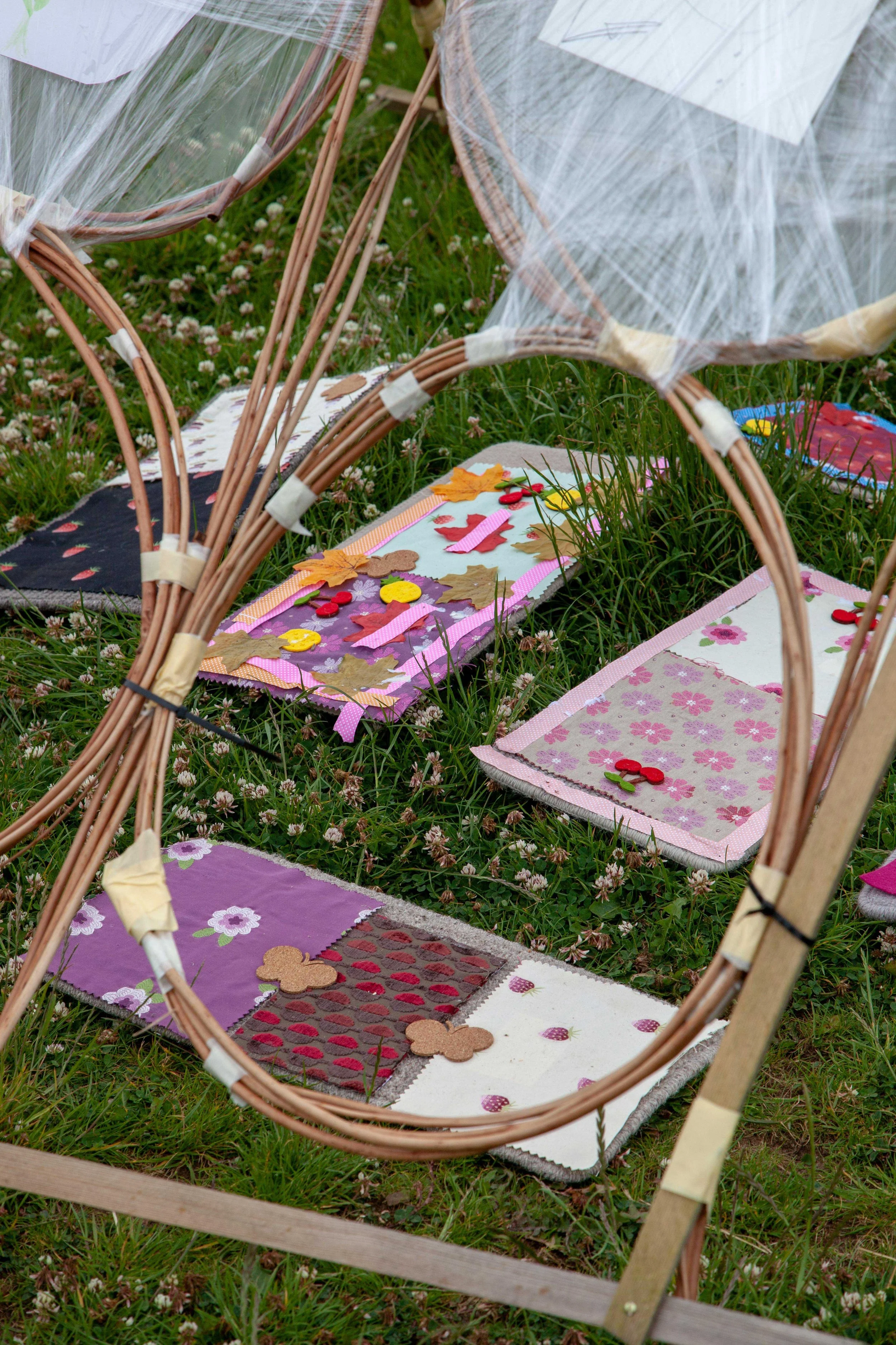 Multiple quilted fabric mats with autumn leaves and buttons design, placed on grass, under a semicircular wooden frame structure with plastic sheeting covering part of it.