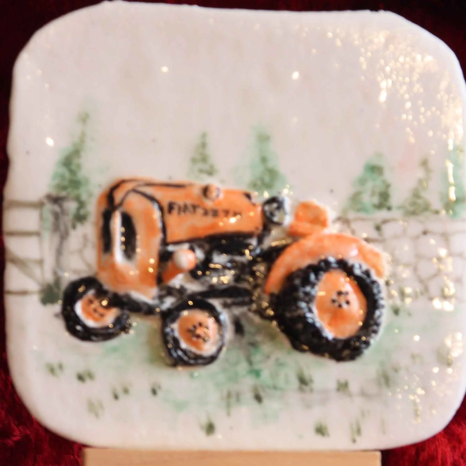 Hand-painted ceramic tile with a winter scene of pine trees and a snow-covered landscape, featuring an orange tractor and black wheels.