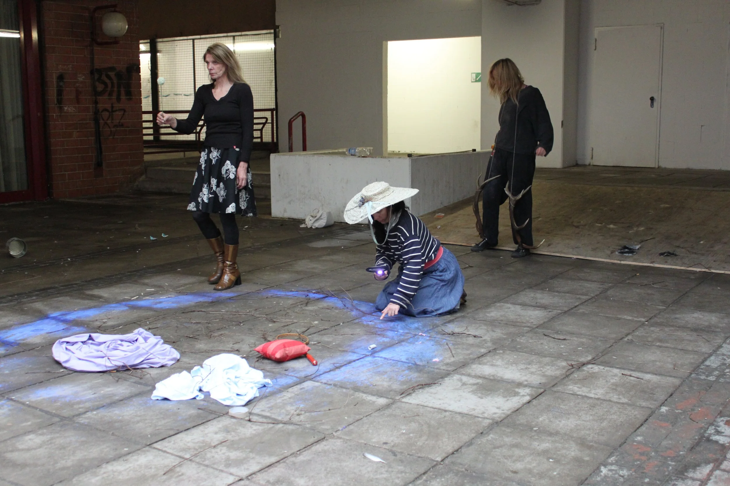 Four women, two standing and two crouching, are in an urban outdoor area with trash and debris on the ground, some connected by electricity or light effects.