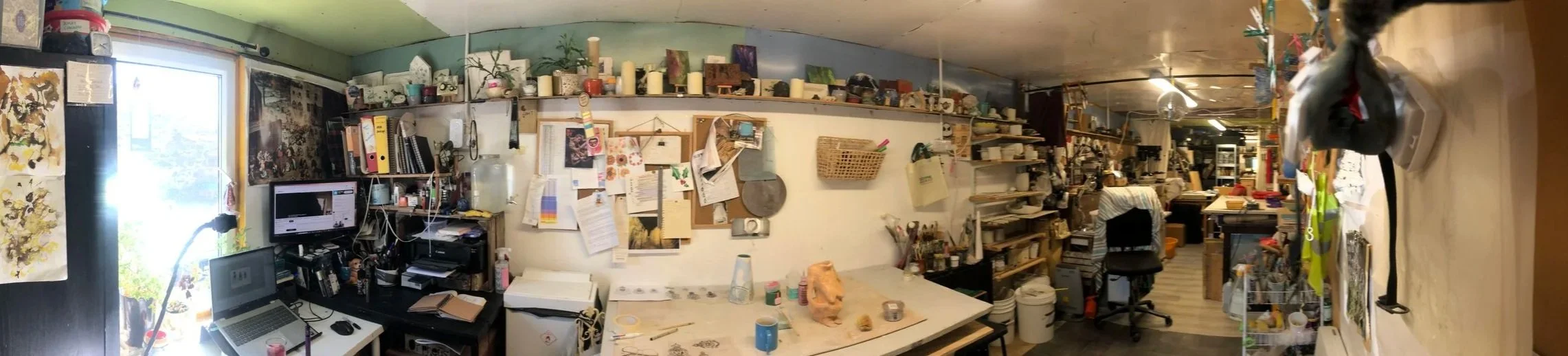 A cluttered art studio with shelves holding various art supplies and decorations, a table with clay and tools, a computer, and artwork on the walls.