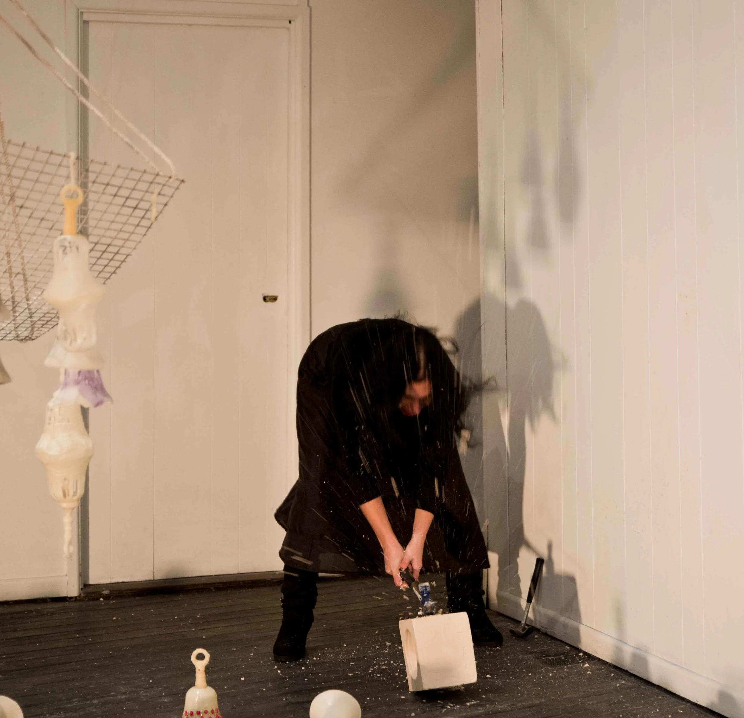 Person in dark clothing smashing objects with a hammer in a room with white walls, broken items, and scattered debris on the floor.