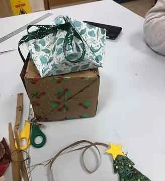 Two wrapped gift boxes, one with a green and white ribbon bow, on a table with craft supplies and a small decorated Christmas tree