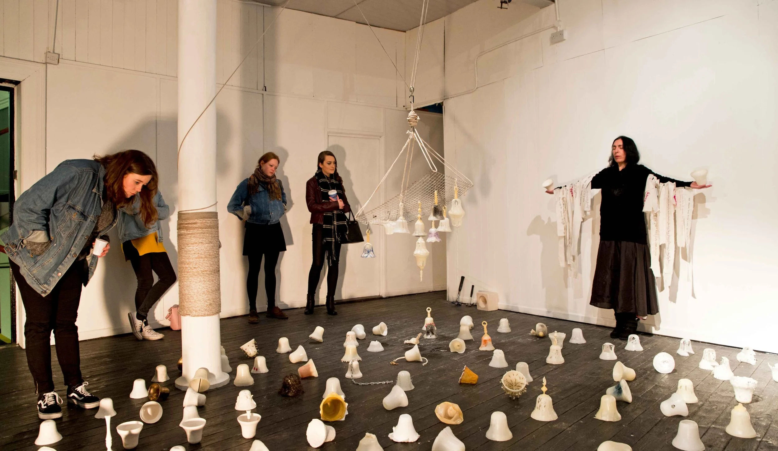 An art installation with various ceramic cups and bell-shaped objects scattered on a dark wooden floor. Four women observe the display, and one appears to be explaining or guiding. The environment is an indoor gallery with white walls and minimal dec