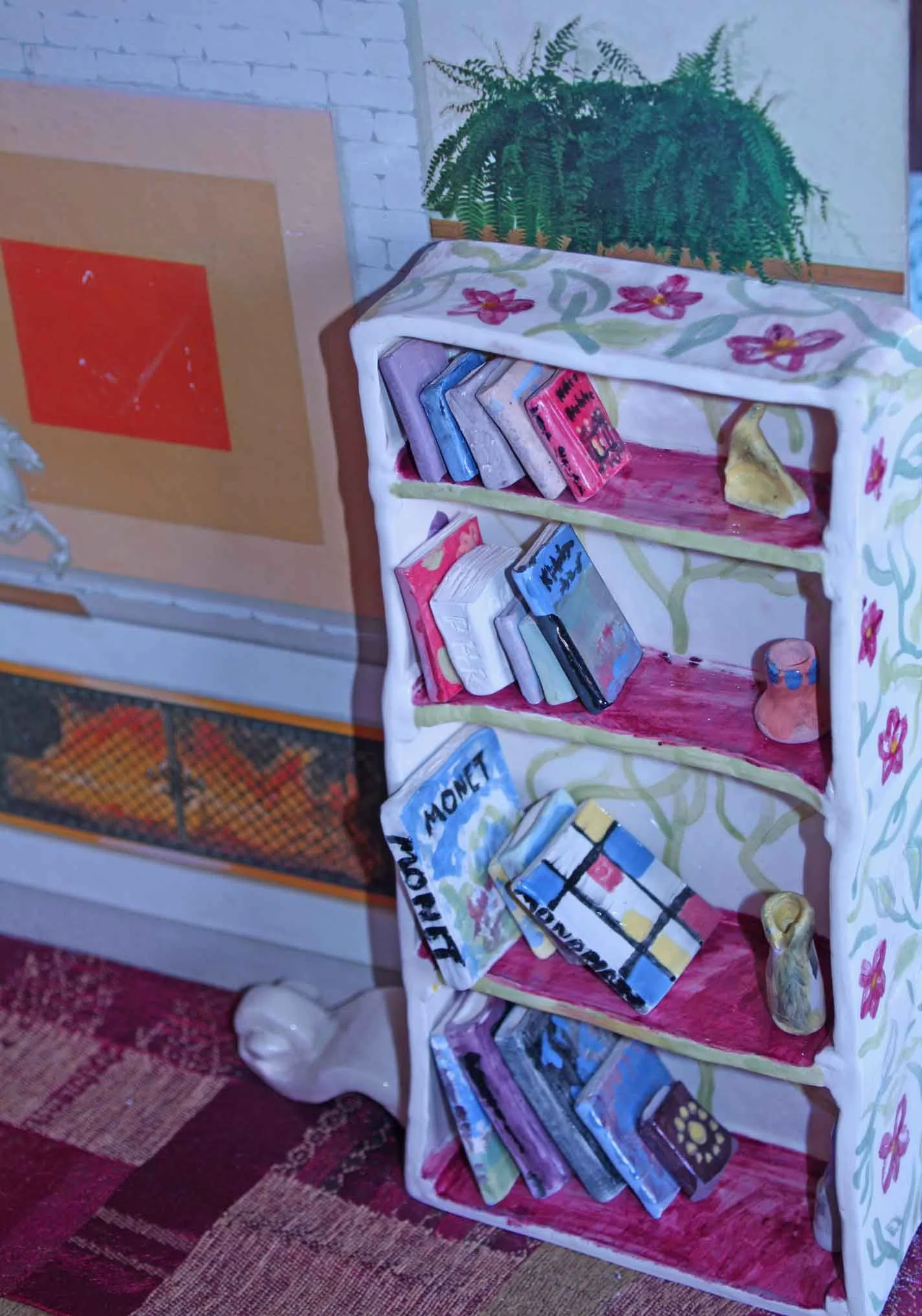 Painted wooden bookshelf with books, a cup, and decorative items on a floral tablecloth, next to a green potted fern and a fireplace.