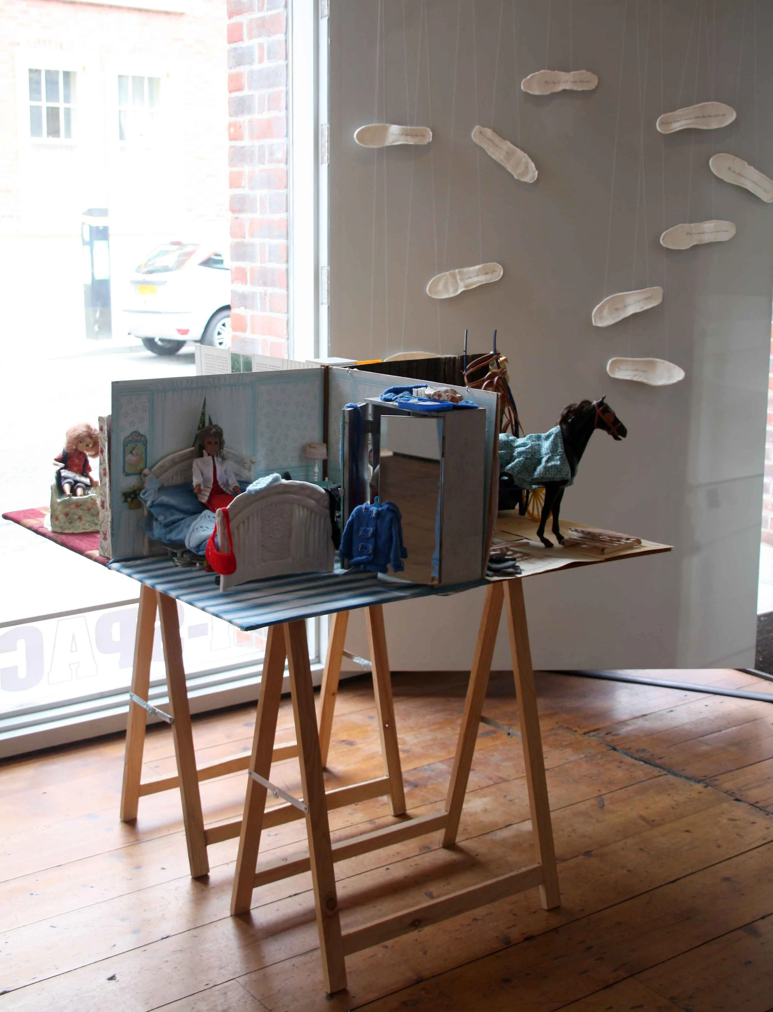 A display of dolls and toy horses inside a store with a large window showing parked cars outside. The display features dolls in beds and toy horses, arranged on tables and the floor with a wooden floor and a brick wall visible.