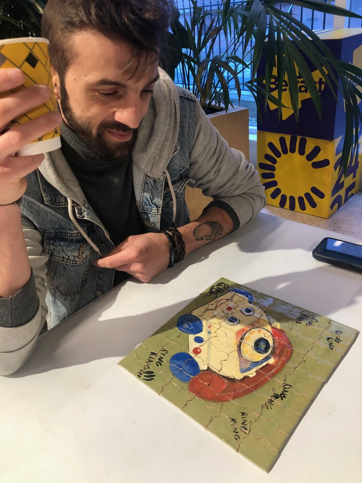 A man with a beard and tattoos is sitting at a white table, holding a yellow paper cup. There is a colorful puzzle on the table depicting a cartoonish face, possibly inspired by a painting or a character. In the background, there are large potted pla