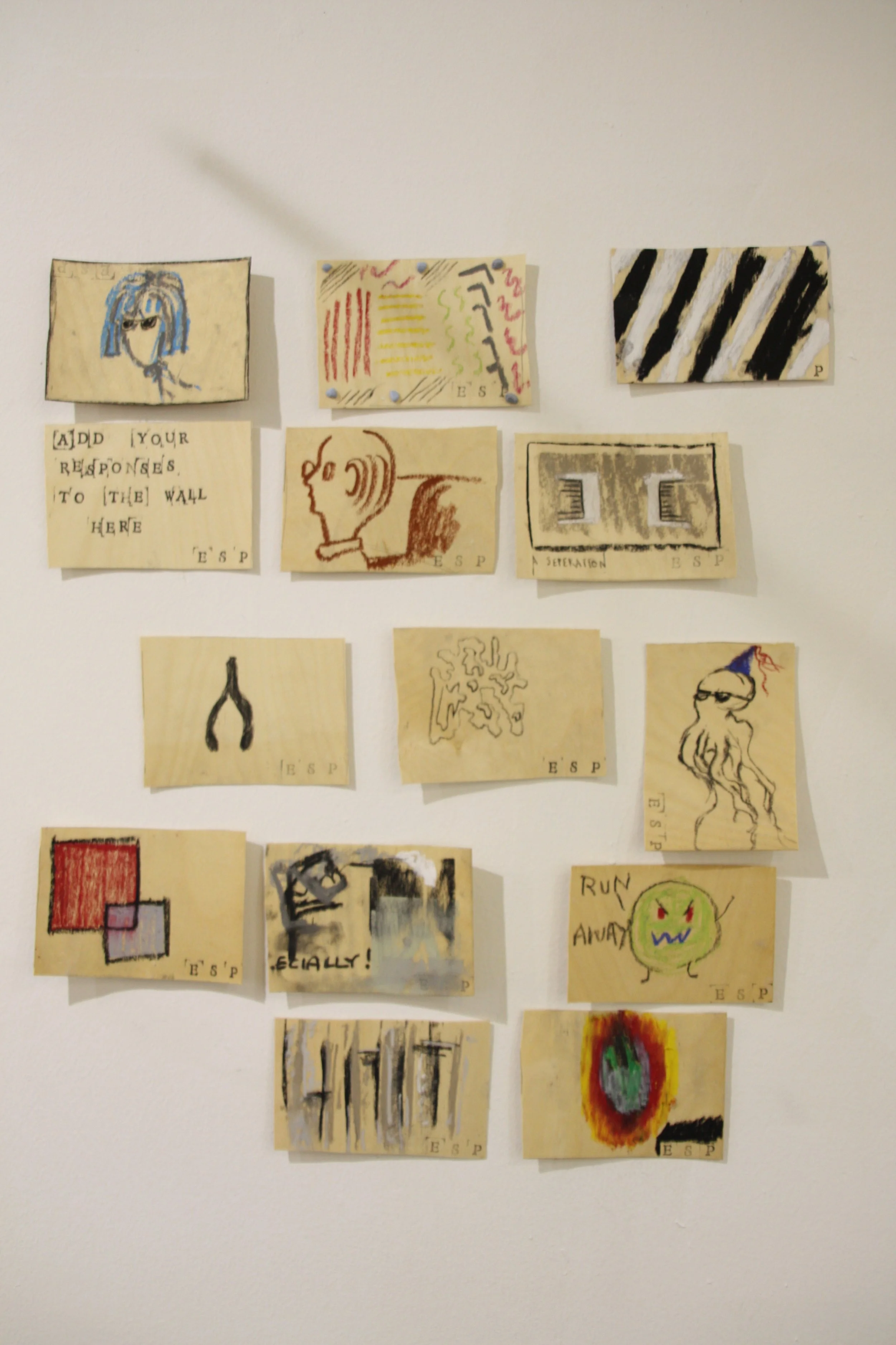 Collection of small, hand-drawn art pieces attached to a white wall. The artworks include abstract designs, faces, and geometric shapes, many with bold lines and colorful detailing.