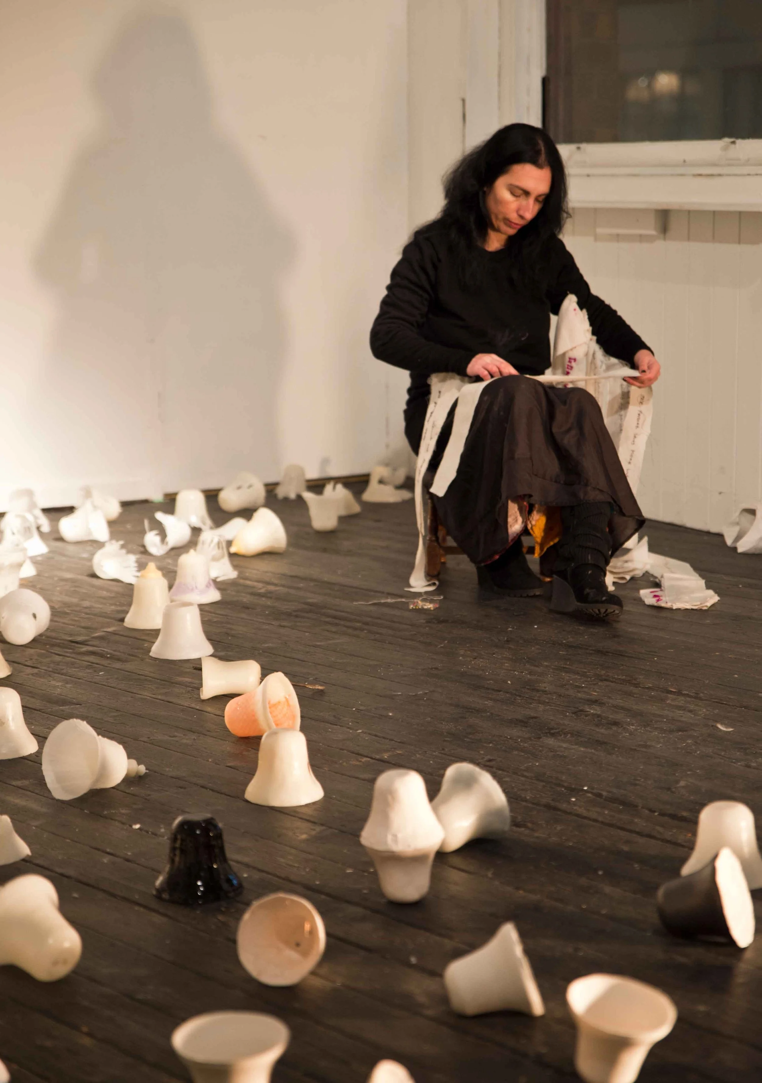 A woman sitting on a chair reading a book, surrounded by scattered seashells on a wooden floor in a room with a white wall and window.