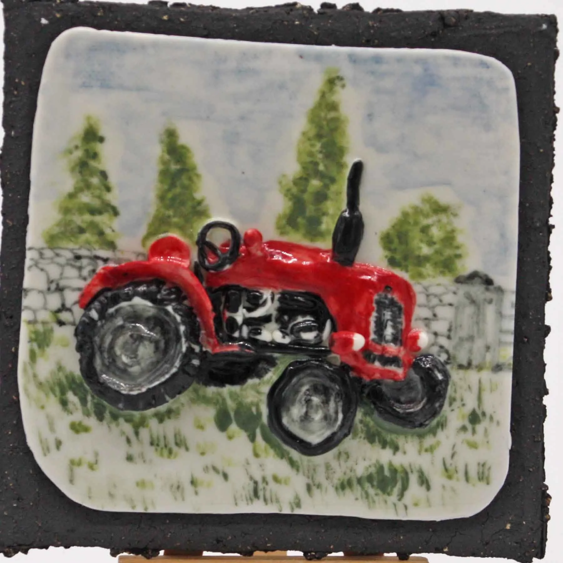 A decorated cake designed to look like a red tractor with black wheels, and a fondant backdrop featuring painted green trees and a cloudy sky.