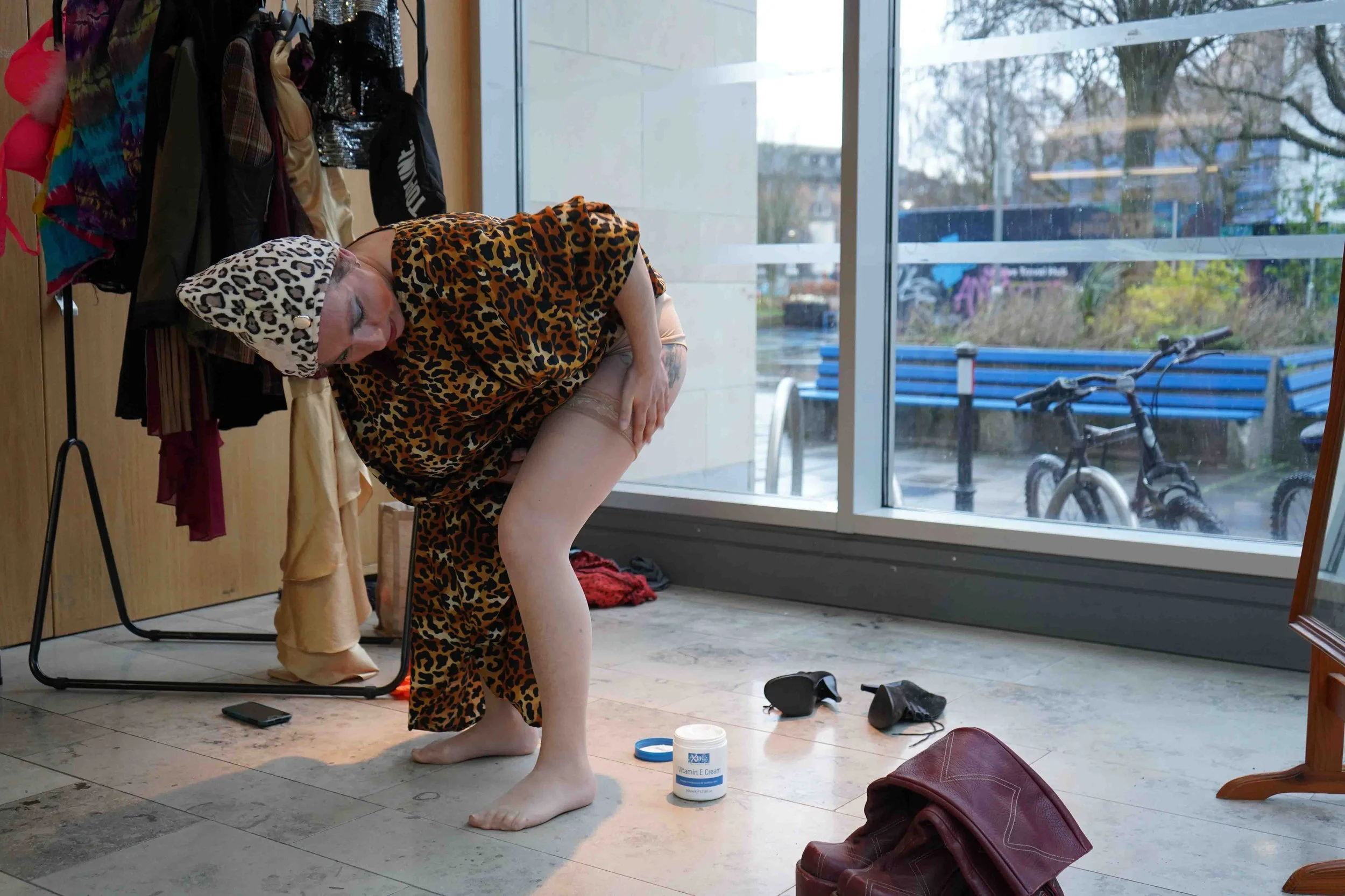 A woman with a leopard print headscarf and dress is posing while holding her leg. She is inside a room with a glass window, a coat rack with clothing, and various personal items on the floor, including a container of Vitamin E cream, shoes, and a wal