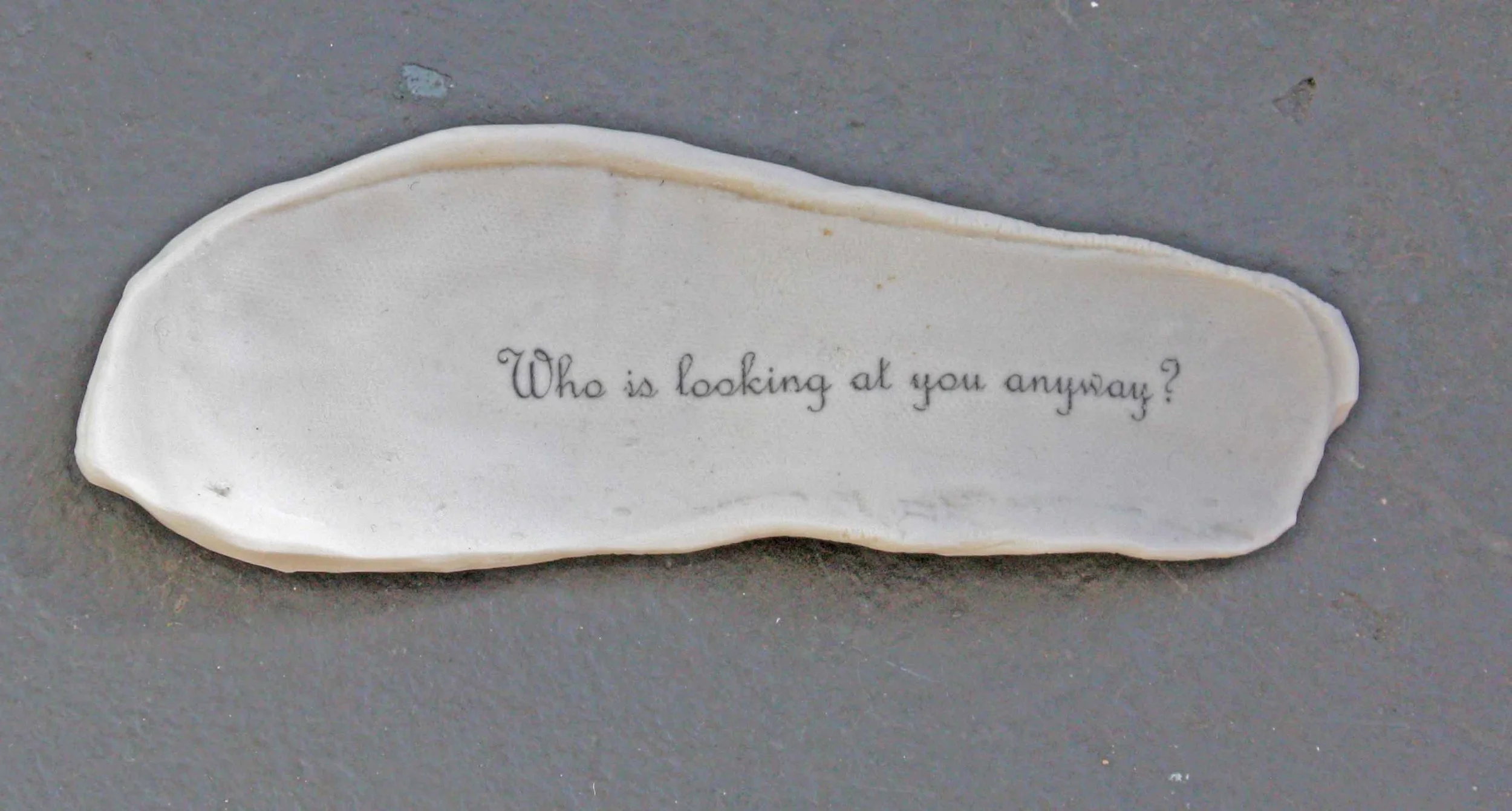 A white ceramic footprint with the engraved words 'Who is looking at you anyway?' on the bowl, resting on a gray surface.