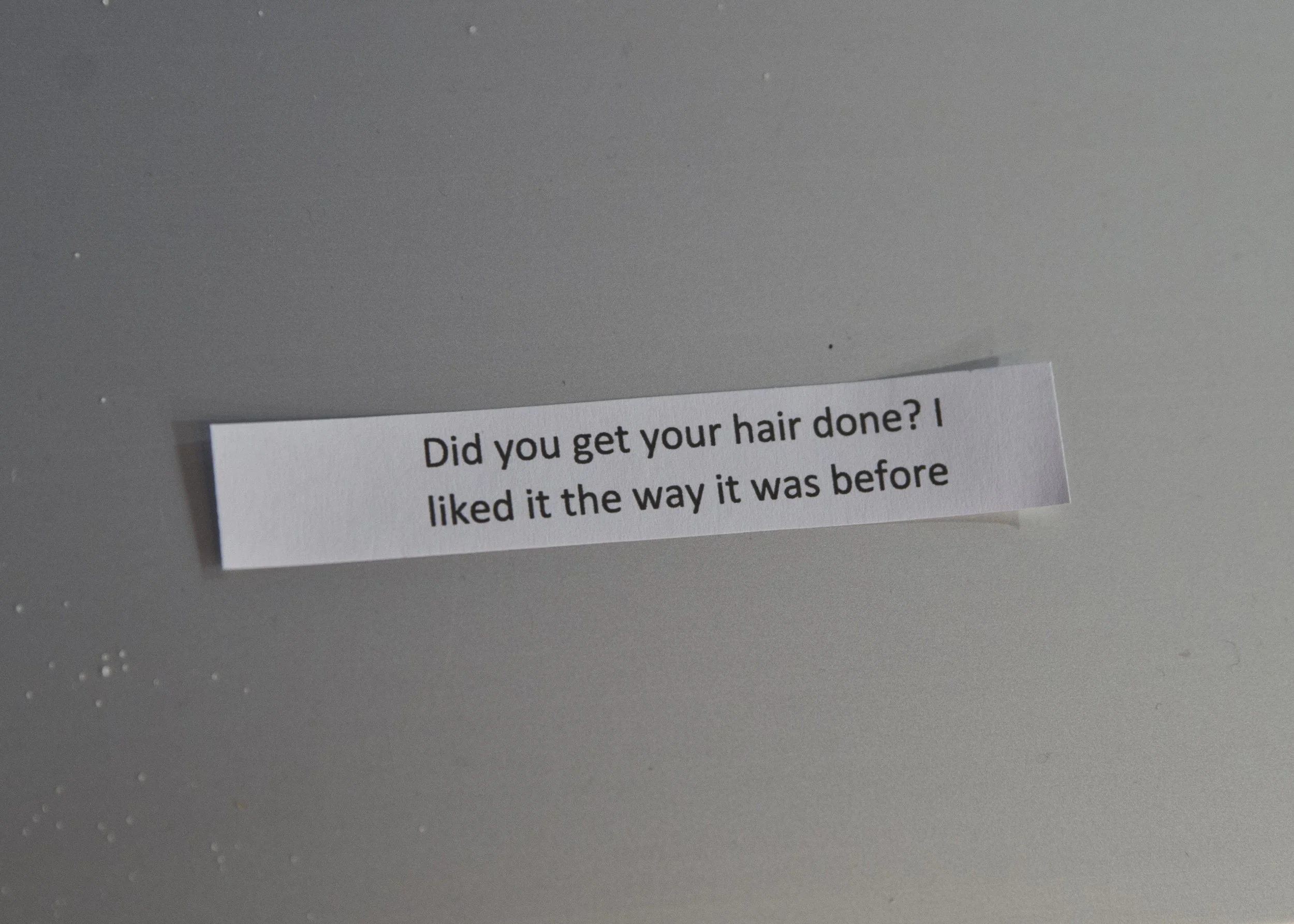 Close-up of a piece of paper with text on it, placed on a surface. The visible part of the text reads: "Did you get your hair done? I liked it the way it was before".