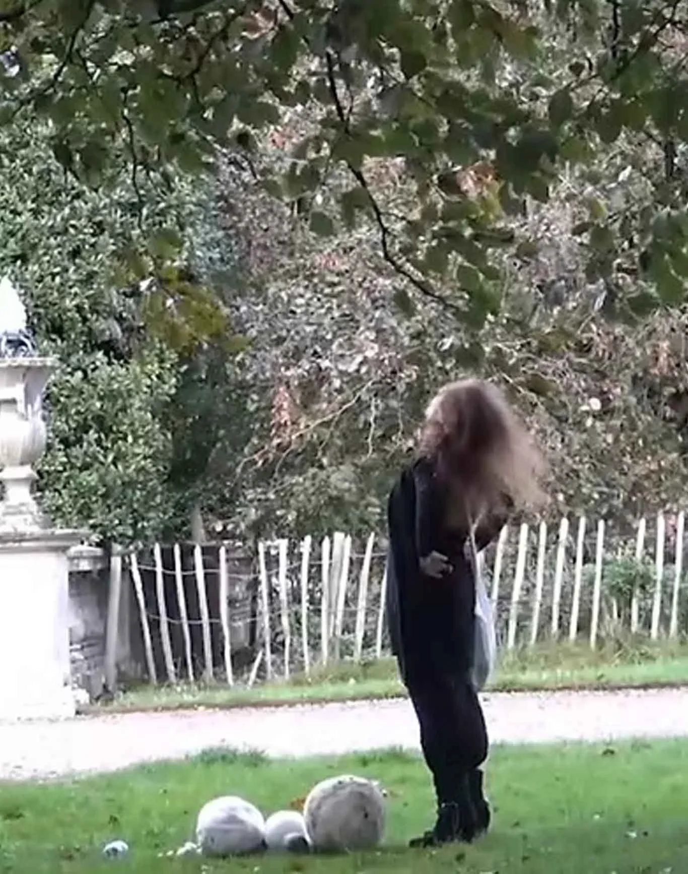 A person with long hair, dressed in dark clothing, standing outdoors near a white wooden fence, with trees and a building in the background.