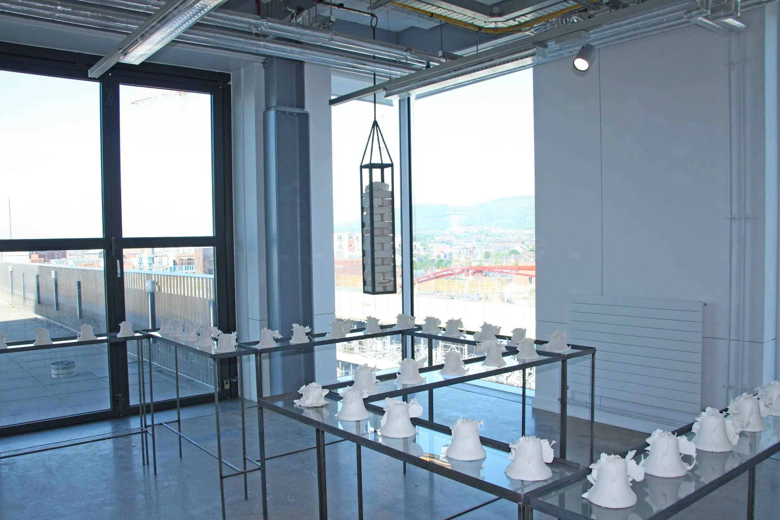 Interior view of a modern building with large windows, construction scaffolding, and numerous white sculptures on display, including chess piece-shaped sculptures and abstract forms.