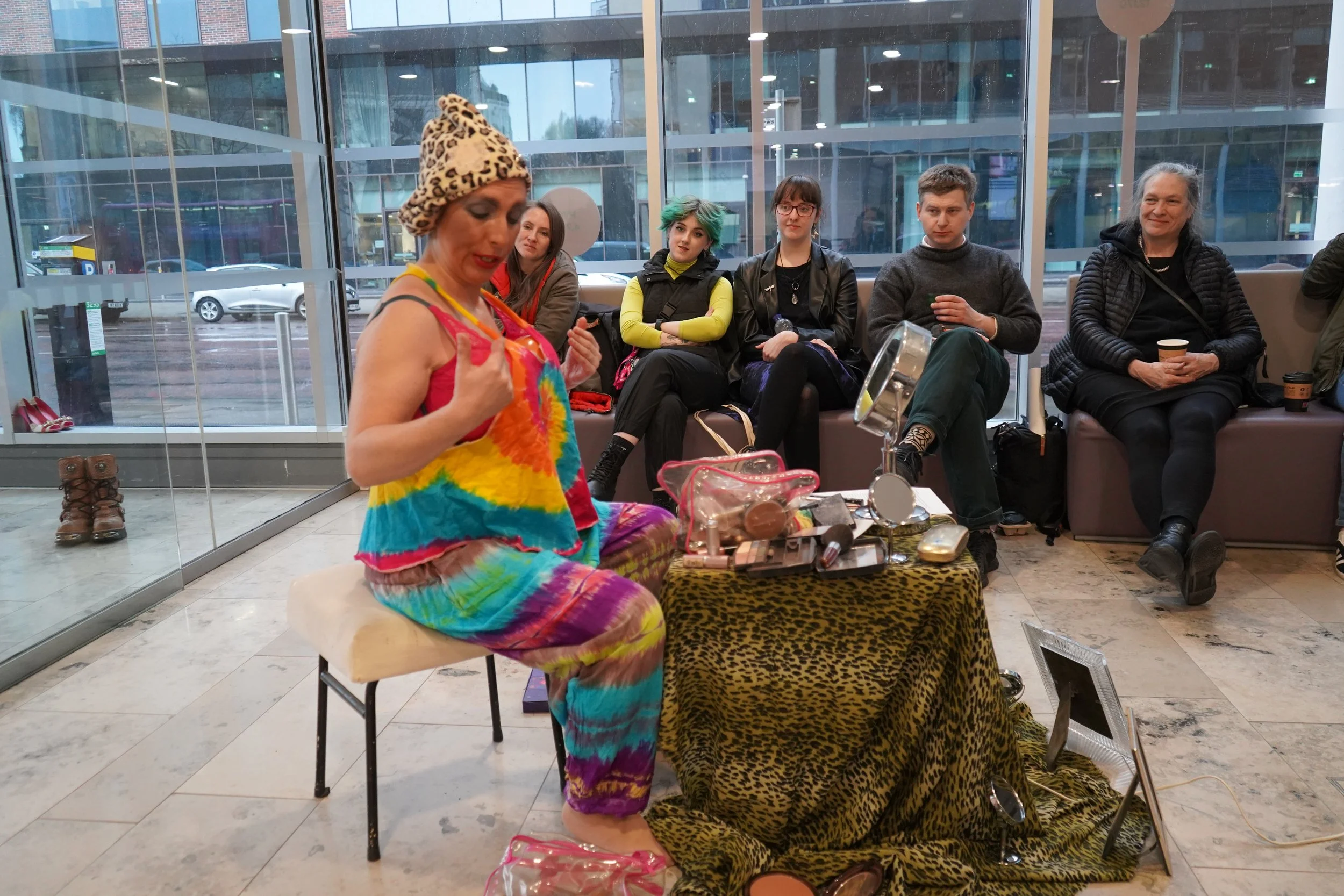 A woman wearing a colorful tie-dye dress and a leopard print hat appears to be performing or speaking in front of a seated audience at an indoor location with large glass windows. Six people are sitting on a bench, watching her, with some holding cup