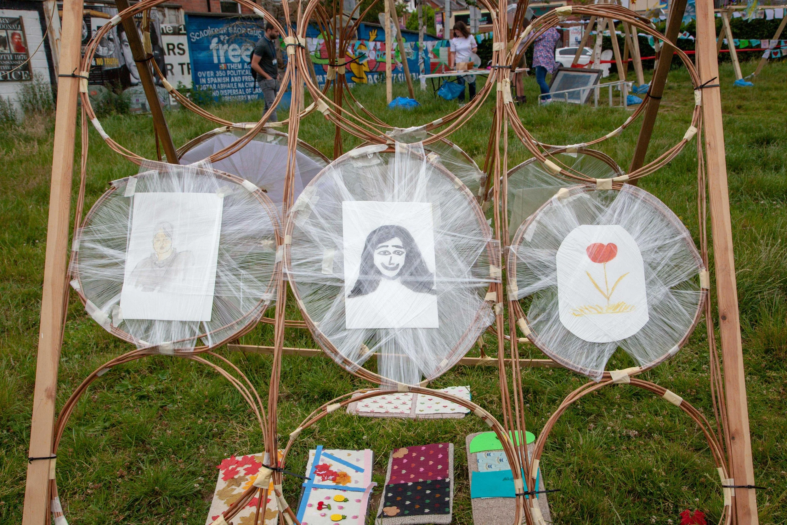 Outdoor art display featuring framed illustrations of faces and a flower, with fabric and felt crafts in various colors and patterns arranged on a wooden frame, at a festival or market.