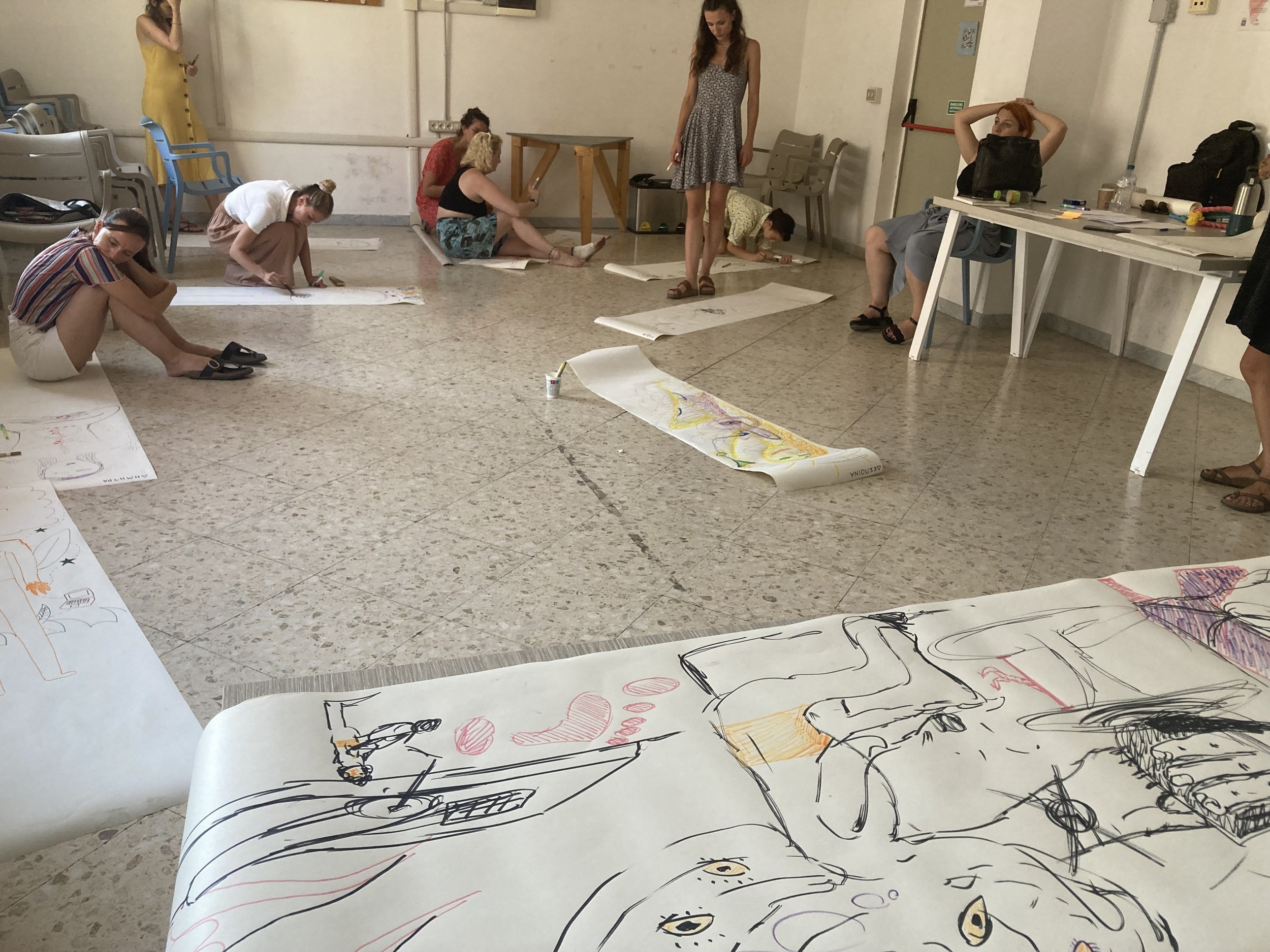 Several people are sitting or lying on the floor in an art studio, drawing on large sheets of paper with colorful markers. Some are focused on their artwork, while others are resting or looking at their phones. There are chairs, backpacks, and art supplies around the room.