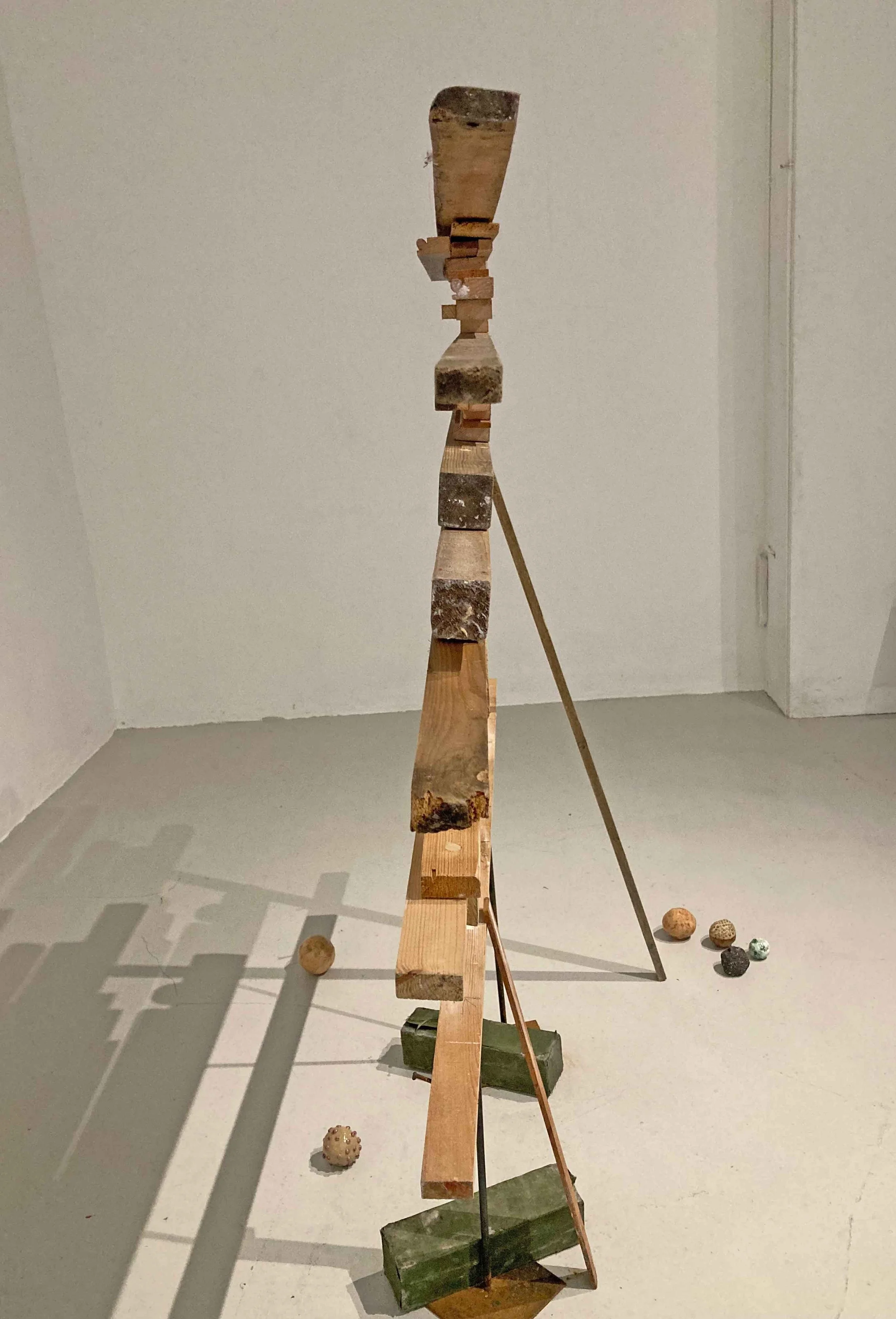An abstract sculpture assembled from various wooden pieces and rocks in an art gallery, with small decorative stones on the floor nearby.