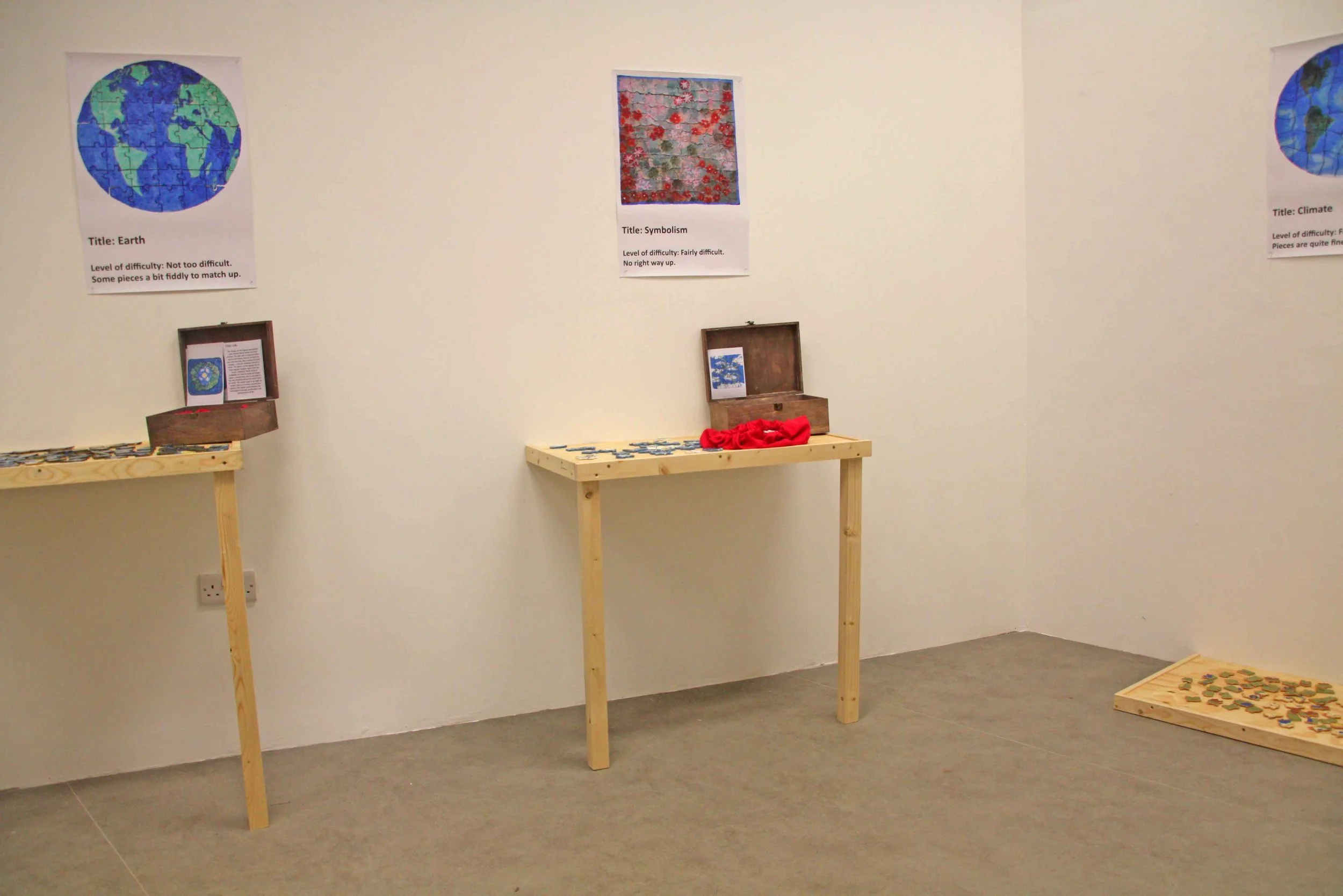 Art display with three posters on the wall, each showing a puzzle image with titles and difficulty levels, and wooden tables with scattered puzzle pieces and open puzzle boxes.