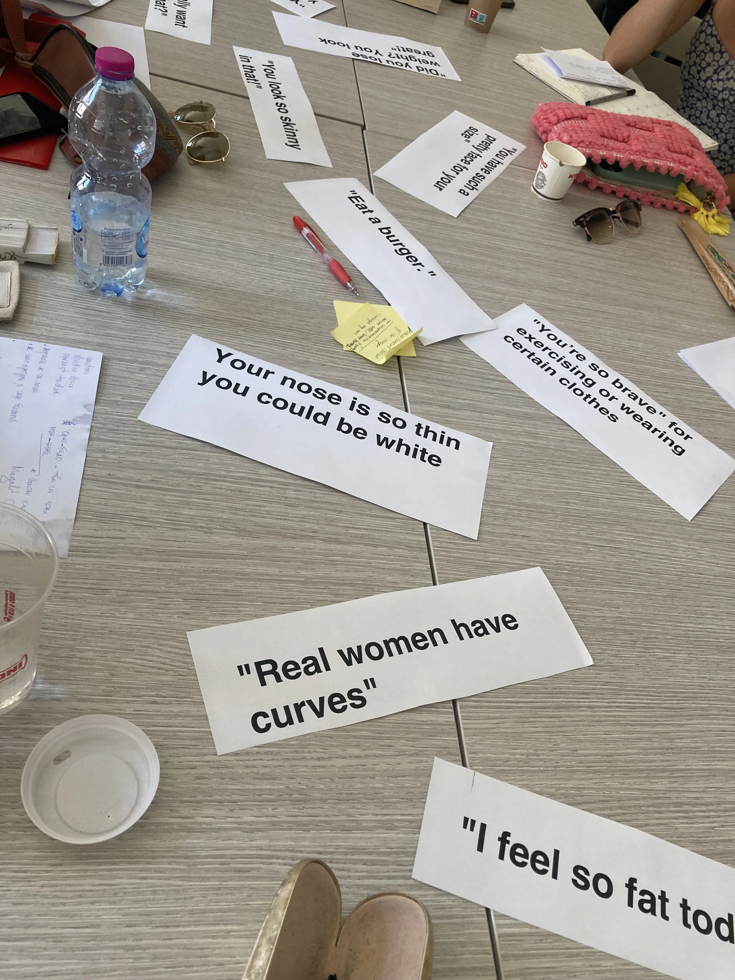 A cluttered table with papers containing quotes about body image and self-esteem, a water bottle, sunglasses, a pink knitted pouch, a small paper cup, a pen, and some notes.