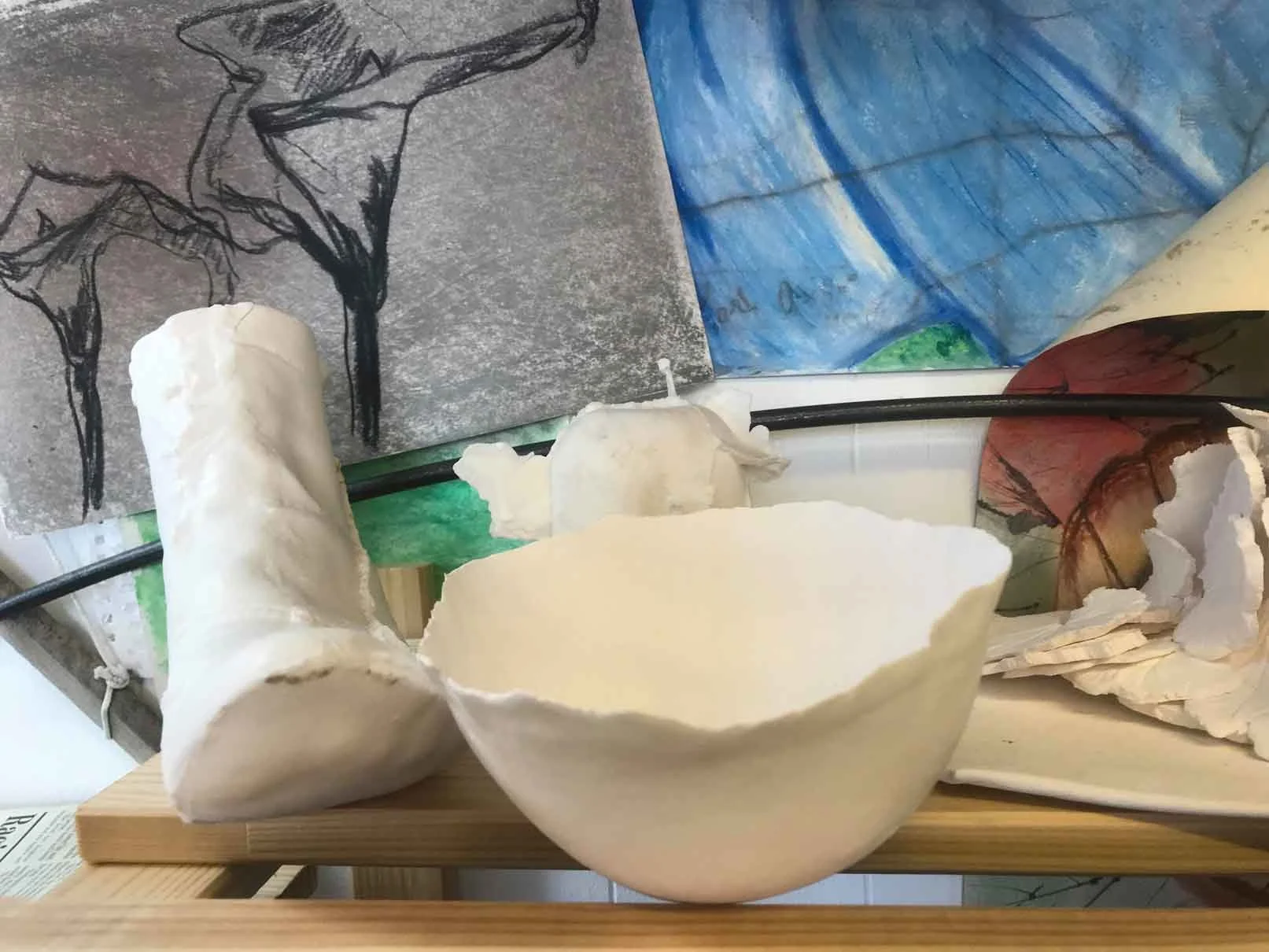 Broken white ceramic cup on a wooden board, with crumpled pieces of paper and artwork in the background.