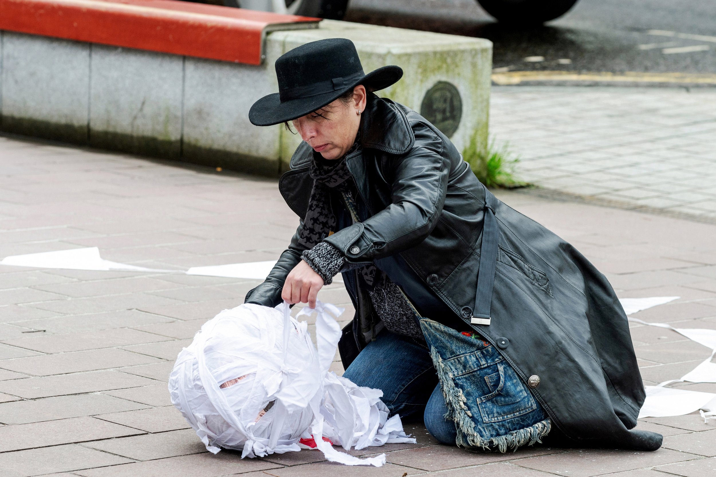 Woman dressed in black leather jacket and wide-brim black hat kneeling on a city sidewalk, with a large ball covered in white fabric.