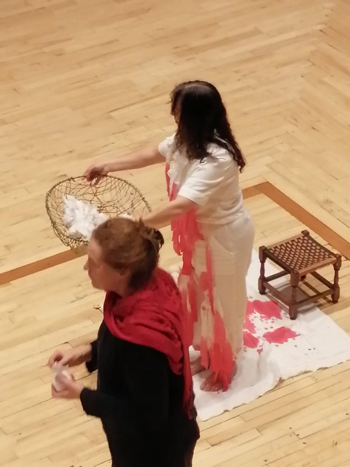 Two women with a third person participating in a ritual or spiritual ceremony on a wooden floor. One woman is sitting on the floor, and the other is standing, holding a basket with white cloth or paper inside. There is a small wooden stool and white 