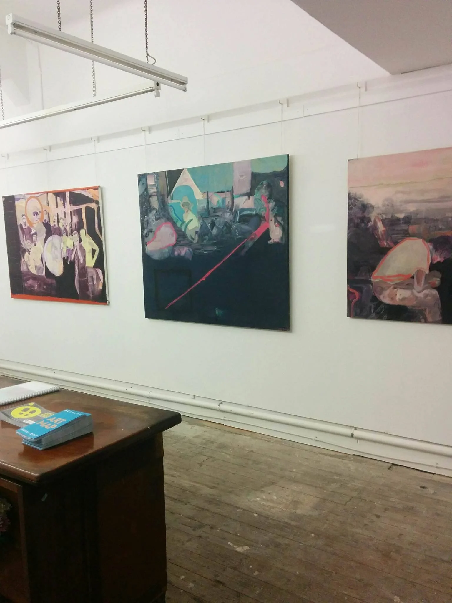 Art gallery displaying abstract paintings on a white wall, wooden floor, table with brochures in the foreground.