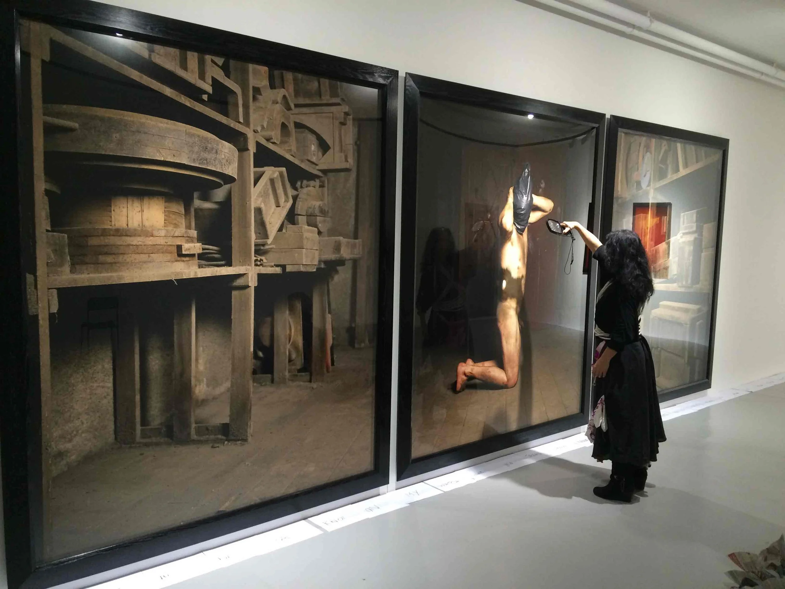 A woman and a young girl looking at a large framed artwork in a museum. The artwork depicts a partly nude person with some black fabric on their head, standing with their hands behind their head. The scene is slightly reflective on the glass covering