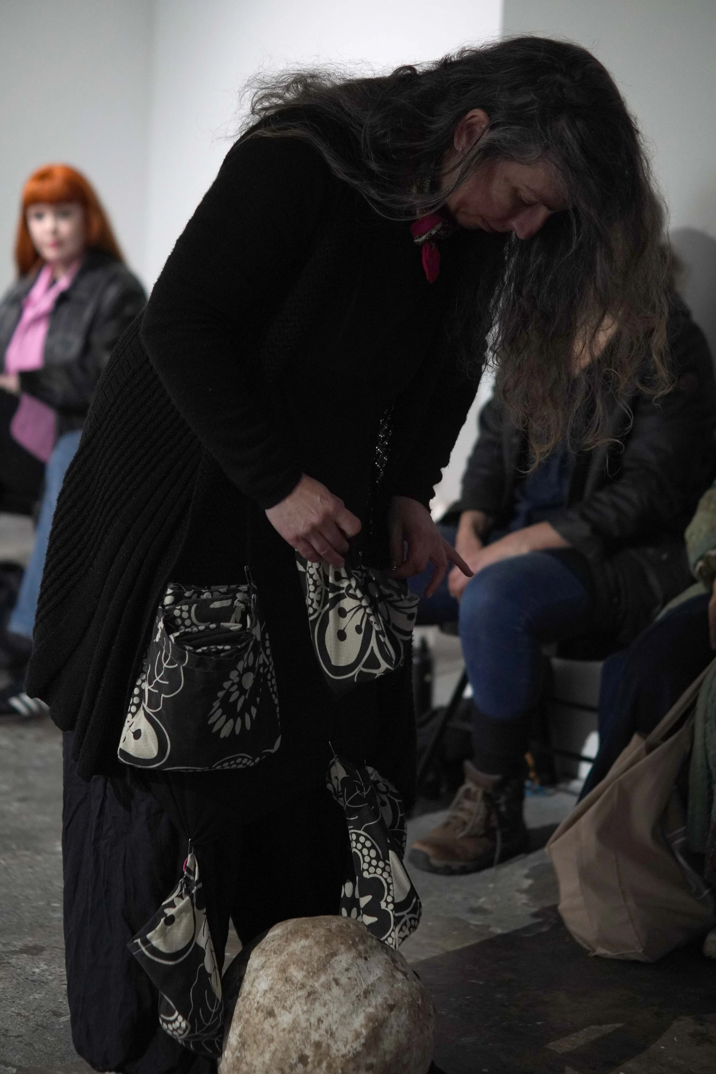 A woman in black clothing bending over a large rock, with a patterned bag hanging from her shoulder, at an indoor event.