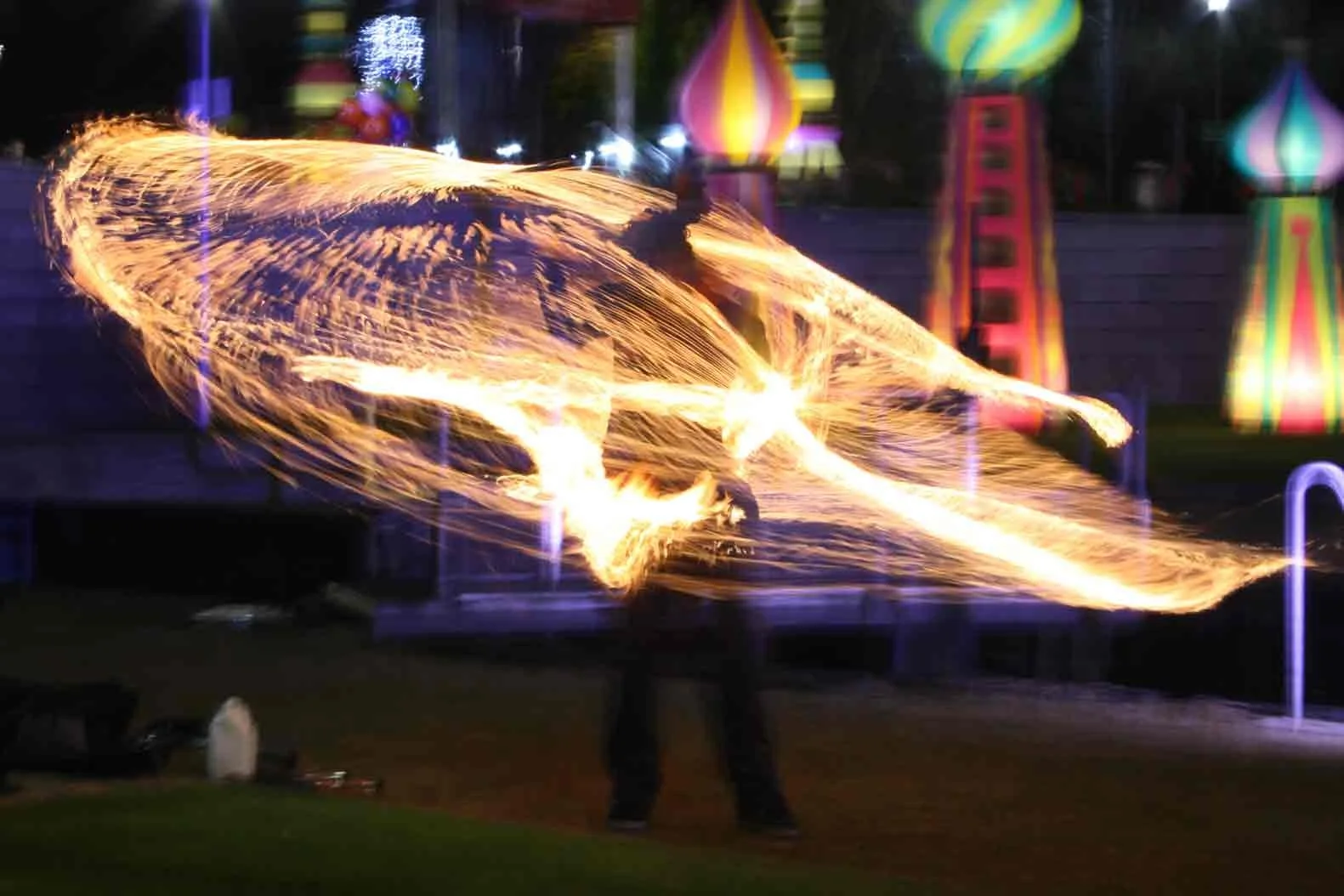 Person performing fire spinning or fire dancing outdoors at night, with colorful illuminated structures in the background.