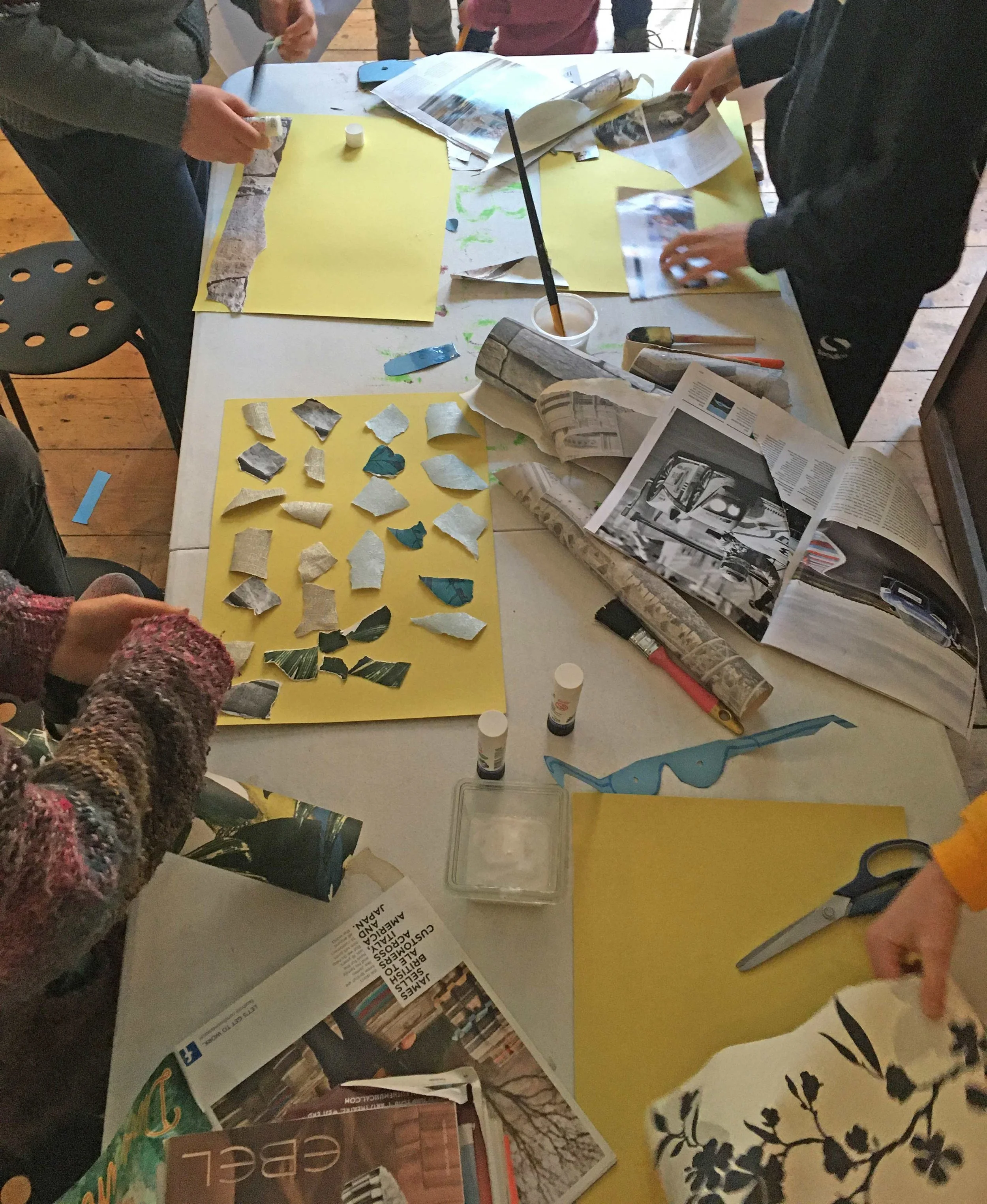 A group of people gathered around a table working on a project involving torn pieces of paper, glue sticks, magazines, and newspapers. The table is cluttered with various craft supplies and colorful papers.