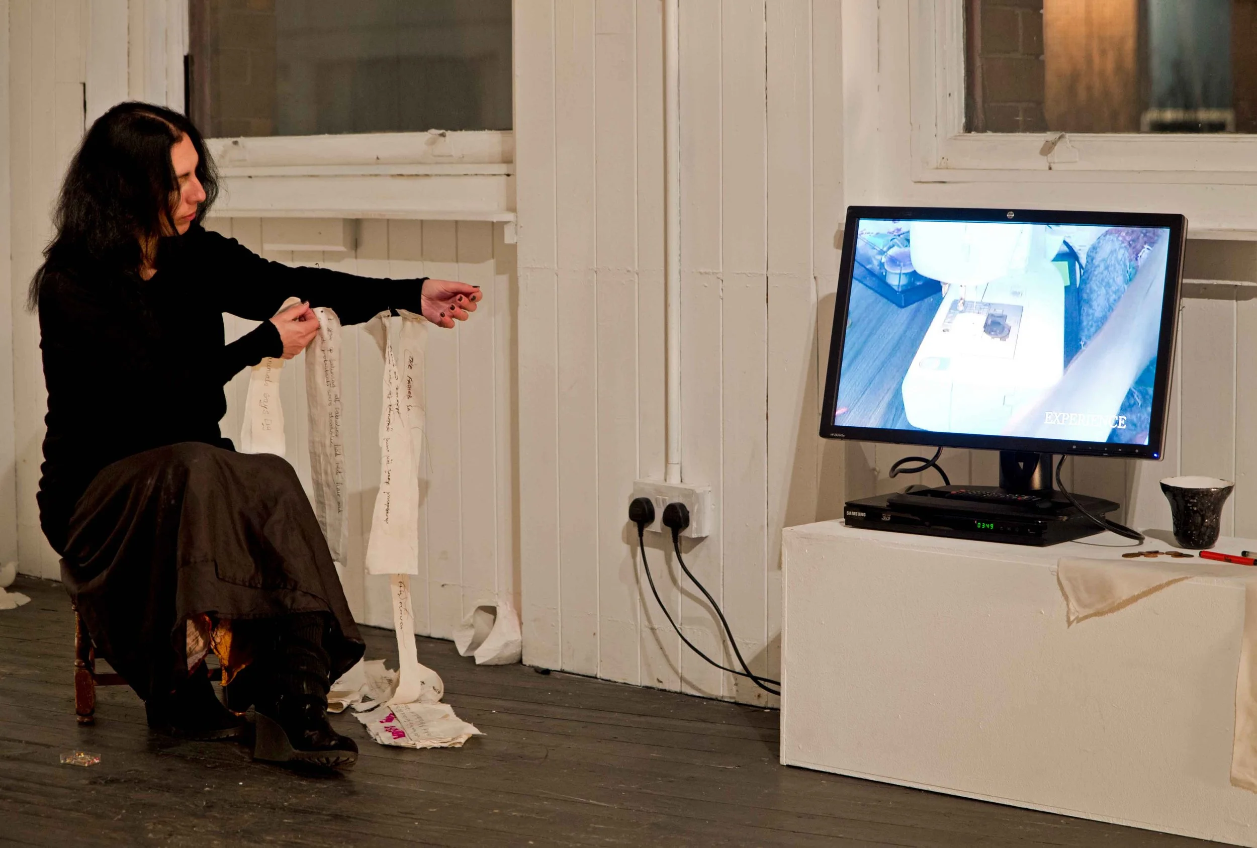 A woman with black hair and a black top is sitting on a chair in an art gallery, examining scraps of handwritten fabric or paper. There is a computer monitor to her right displaying an image of an interior space, with a black device and a cup on a wh